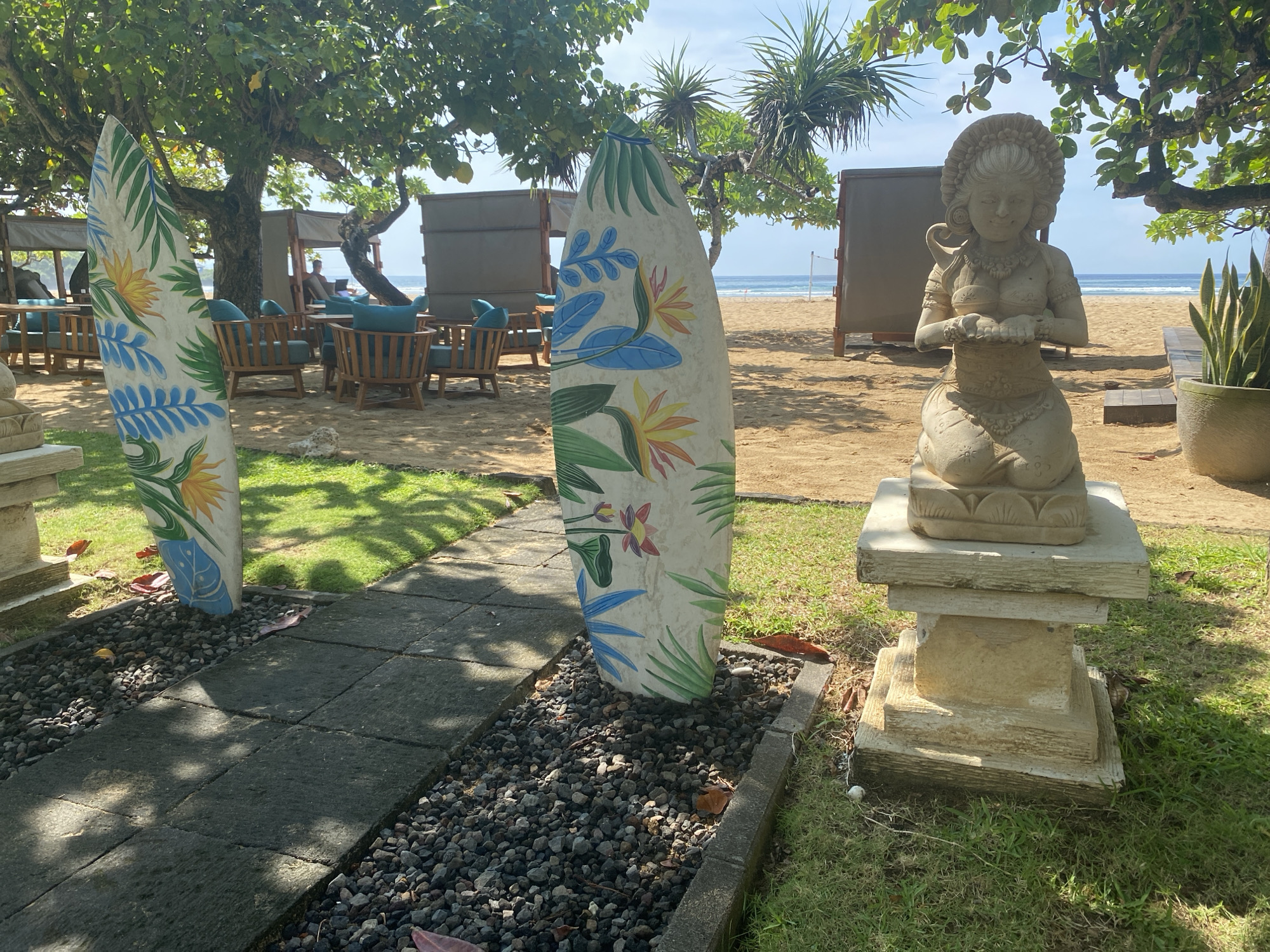 The image captures a serene scene in Nusa Dua, Indonesia, likely near a beach resort or coastal area. In the foreground, two decorative surfboards stand on either side of a stone pathway lined with small dark rocks. The surfboards are painted with floral designs. To the right, a stone statue of a kneeling female figure is placed on a tiered pedestal. The background reveals a sandy beach with lounge chairs under shaded structures. The ocean is visible in the distance, and trees provide shade, creating a relaxed and tropical atmosphere. The scene is well-lit with sunlight.