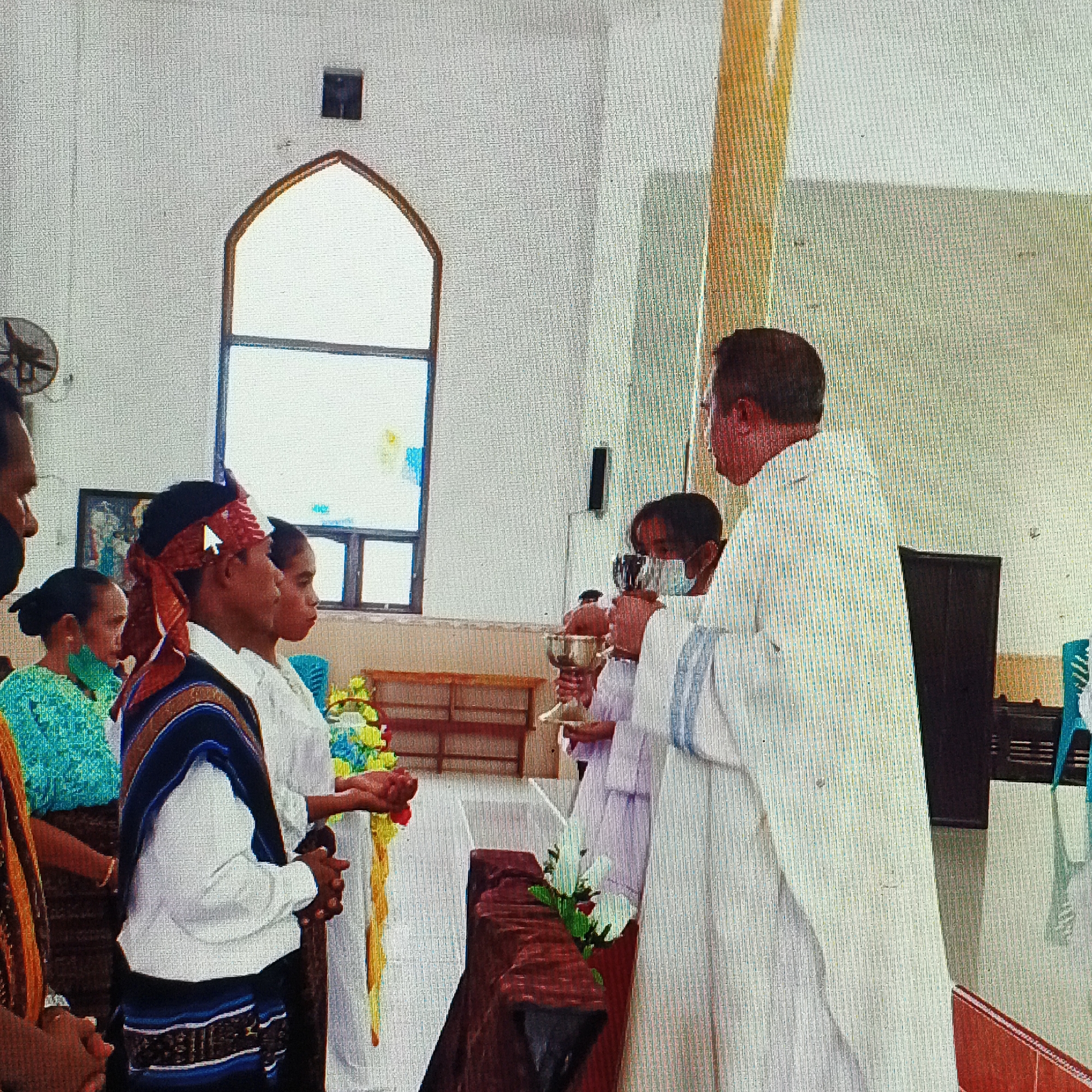 This image depicts a religious ceremony within a church, likely in Ende, Indonesia, judging by the traditional attire of some individuals. A priest, dressed in a white robe, stands on the right side of the frame, holding a chalice and appearing to be administering communion. In front of him are several individuals, including a young man wearing a red and patterned headscarf and a traditional woven garment over a white shirt. Another young person, partially obscured, stands next to him, also in white. Further to the left, a woman in a green top is visible. The background shows typical church architecture with a large arched window and wooden pews. The overall atmosphere suggests a formal religious gathering.