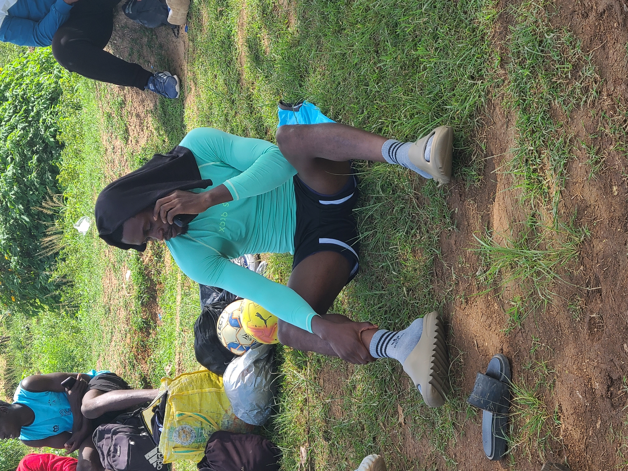 A man wearing a light green long-sleeved shirt, black shorts, and light grey socks with tan sandals is seated on the grass in Unknown, Unknown. He has a black covering over his head. He is holding a phone to his ear. Next to him is a yellow soccer ball, a black plastic bag, and a pair of black sandals. A small group of people sit nearby.