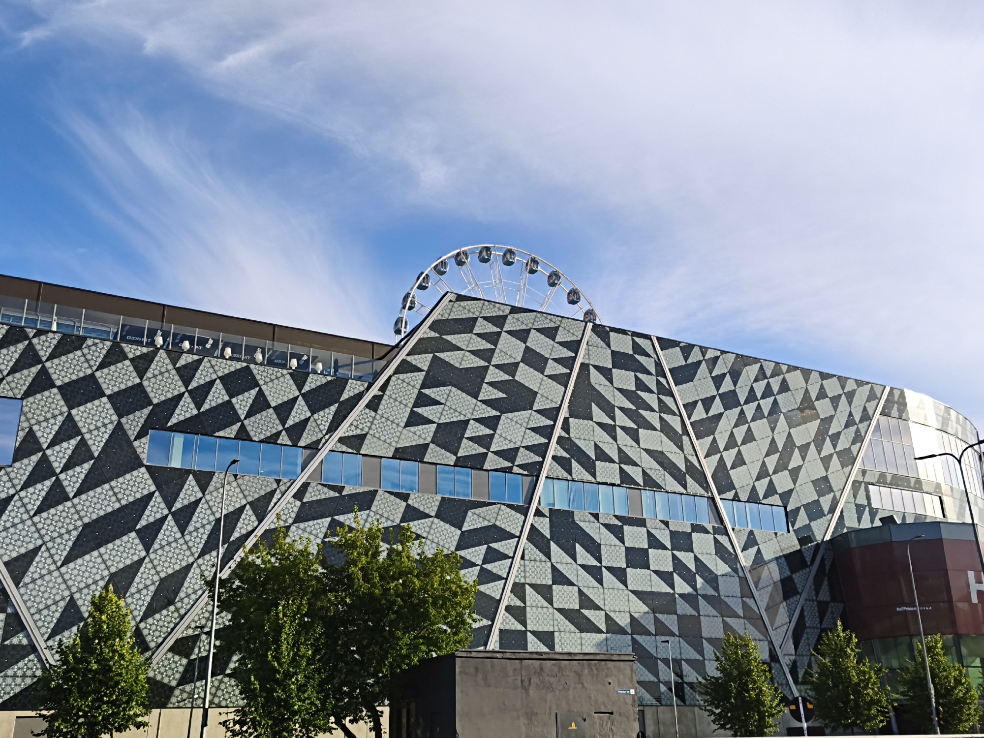 The image shows the exterior of a modern building in Tallinn, Estonia. The building features a distinctive geometric facade composed of dark gray and lighter gray triangular and diamond shapes. A Ferris wheel is visible on top of the building's roof. Several trees are in the foreground, and a street light is positioned in front of the building. The sky above is blue with wispy white clouds. Part of a sign reading "H..." is visible on the right side of the building.