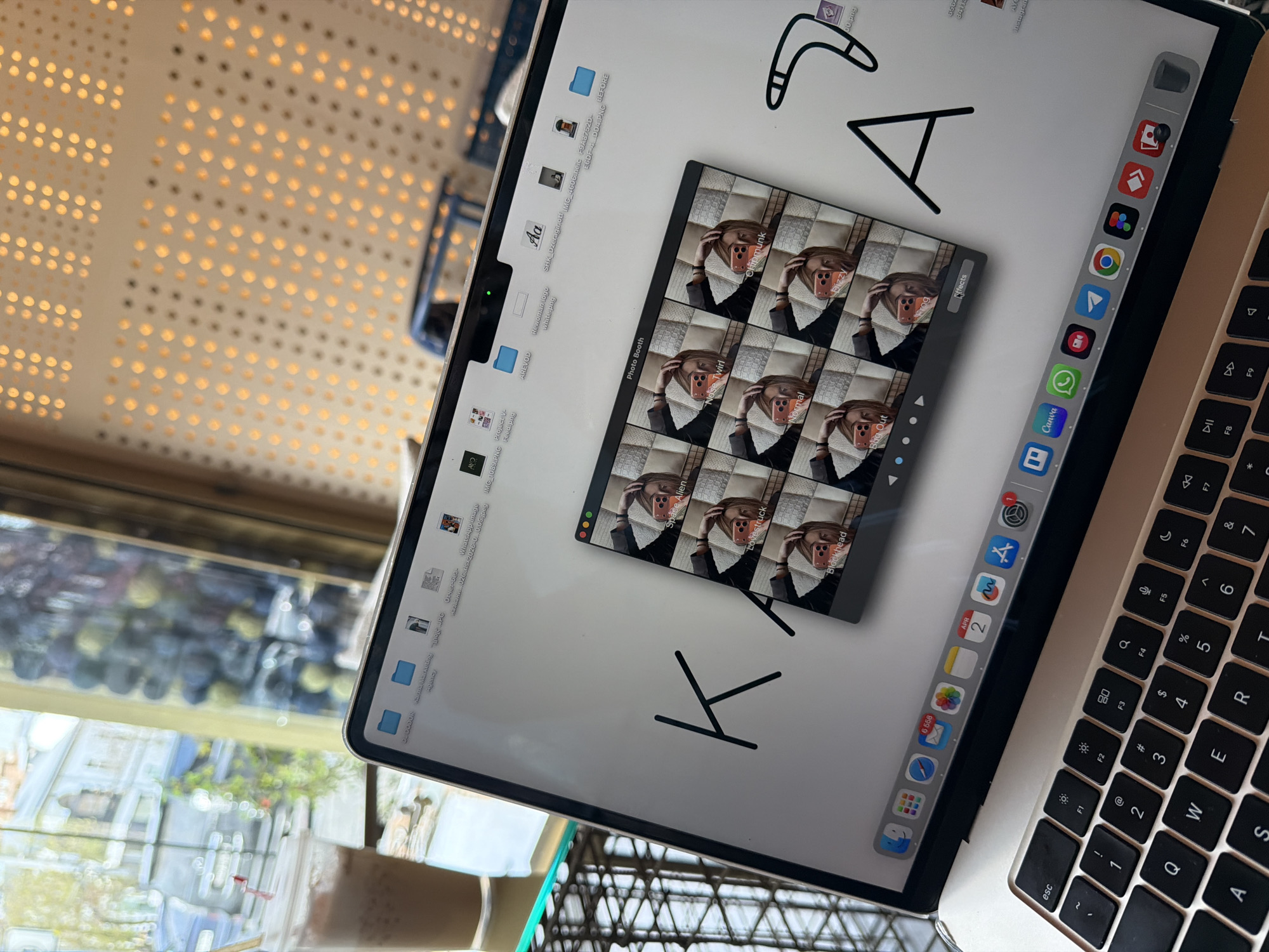 The image shows a laptop on a table, displaying a screen with various icons, folders, and a photo booth application showing different effects applied to a person's image. Handwritten text "AYO KAY" is also visible on the screen. The laptop is partially covering a keyboard. In the background, a blurred scene is visible through a window, showing glimpses of greenery and buildings. Some light fixtures are partially visible in the top left corner. The city of Yerevan, Armenia, is not discernible from the image alone.
