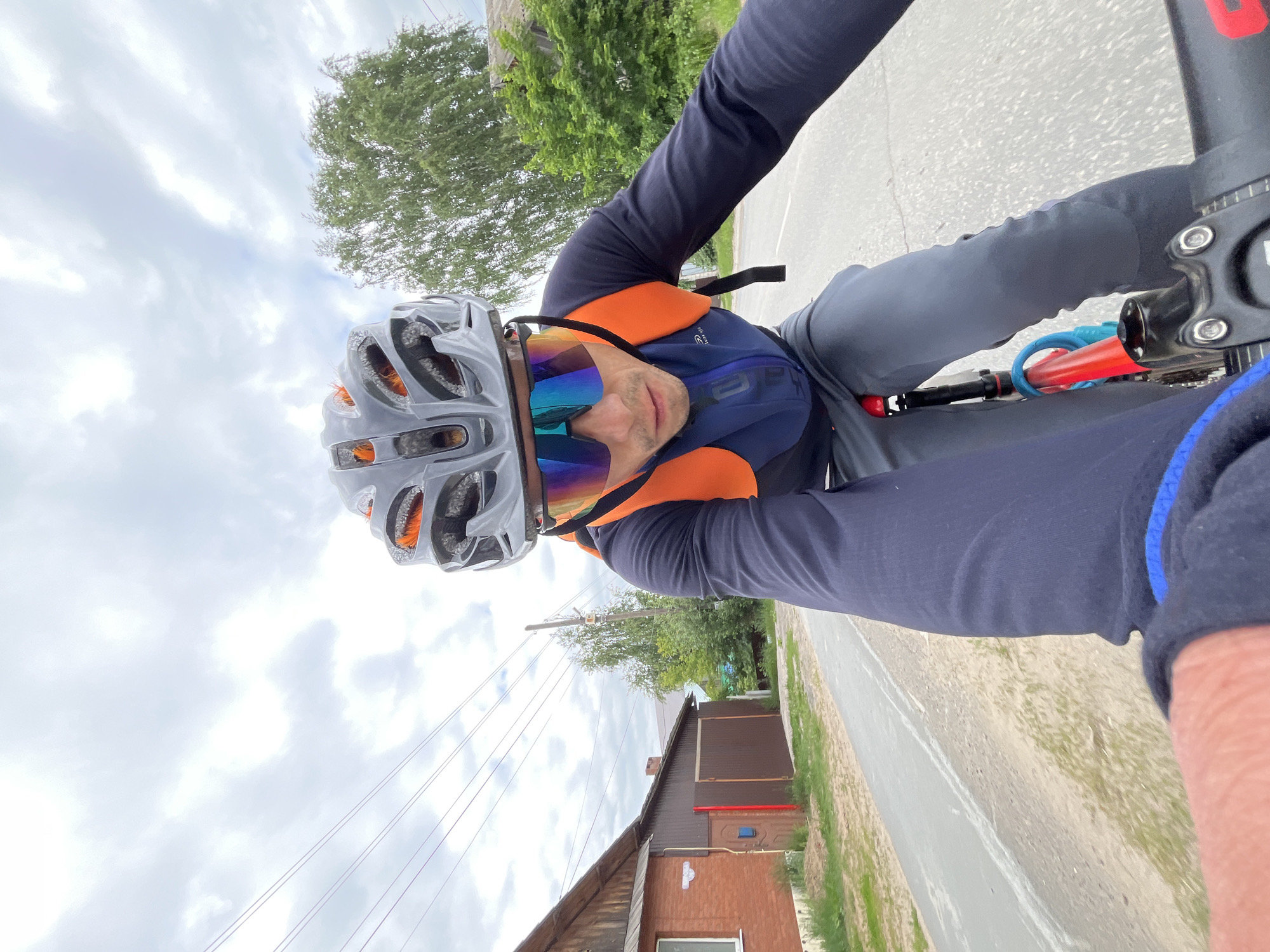 A first-person perspective captures a male cyclist wearing a helmet, sports sunglasses, a blue and orange cycling jacket, and dark pants, as he rides a bicycle on an asphalt road. The cyclist is looking down at the road ahead. In the background, a wooden house with a red brick facade is visible on the left side of the road, along with a green lawn and trees. Power lines stretch across the sky, which is overcast with clouds. The road appears to be in a rural or suburban area on the outskirts of Chaikovsky, Russia, given the presence of residential structures and a seemingly quiet road.