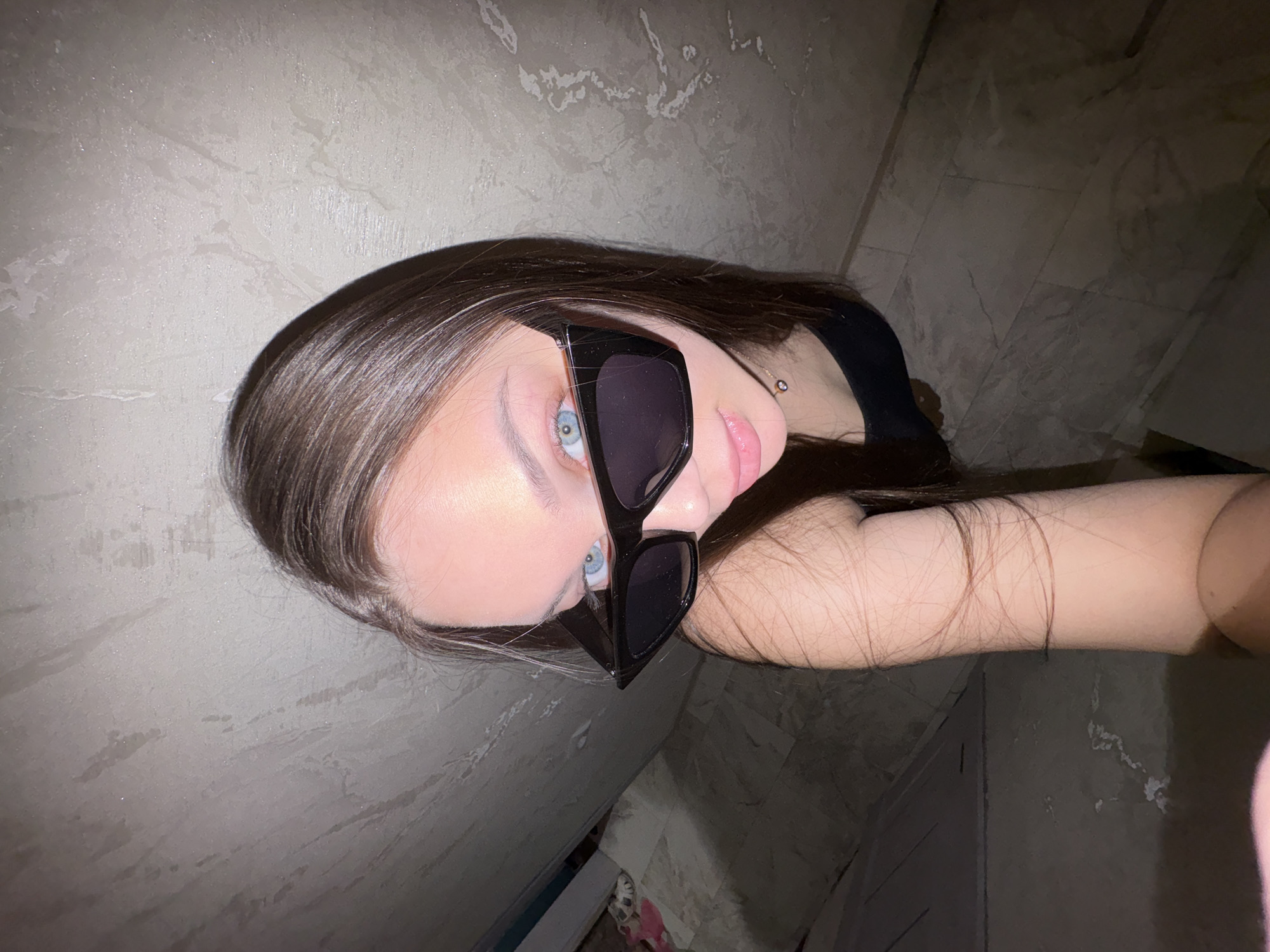 A woman with light skin and brown hair is wearing large, black sunglasses and looking directly at the camera. She is positioned in a room with light-colored, textured walls and dark flooring. The angle of the shot is from below, looking upwards. The location is Chaikovsky, Russia.