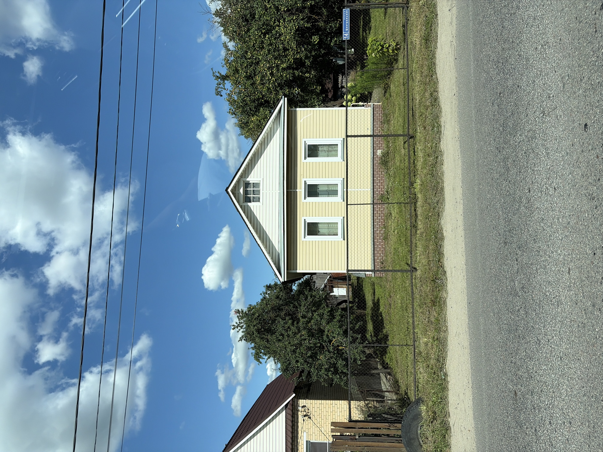 The image depicts a residential scene in the village of Novoye Podvyaznovo, Russia. In the foreground, a paved road with a sandy shoulder runs horizontally across the bottom of the frame. Behind the road, a wire mesh fence encloses a grassy yard.
The focal point of the image is a single-story, light yellow house with white trim, featuring three evenly spaced windows on its front facade. Above the windows, a small gable window is visible. The house appears to be clad in horizontal siding. To the left of this house, a portion of another house with a brown roof and beige brick exterior is visible. A dark-colored tire lies on the grass near the fence on the left side of the yard.
Mature trees with dense green foliage stand behind the houses, creating a natural backdrop. A street sign with Cyrillic lettering, reading "14 Центральная," is affixed to the fence on the right side of the property, indicating the street name and number. The sky above is a bright blue, dotted with fluffy white clouds. Power lines are strung horizontally across the upper portion of the image. The scene is captured during daylight, with sunlight illuminating the houses and yard. There are no people or animals visible in the image.