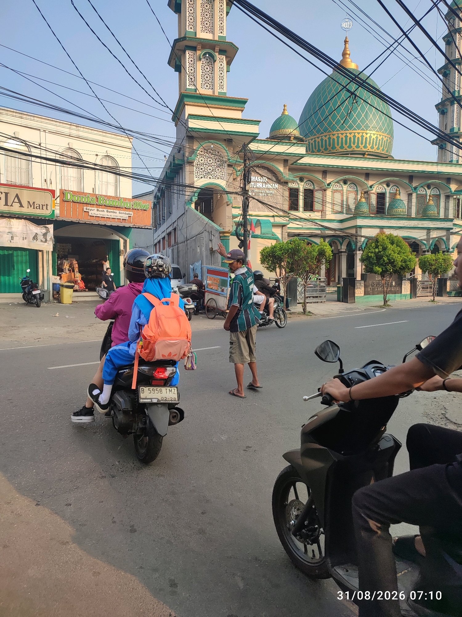 In the city of East Jakarta, Indonesia, on August 31, 2026, at 07:10, a street scene unfolds under a clear sky. The dominant structure is a mosque with a large green dome adorned with gold geometric patterns, flanked by minarets and featuring an arched facade with multiple windows. In front of the mosque, two individuals are seen on a motorcycle. The rider, wearing a pink jacket and a black helmet, is followed by a passenger in a blue headscarf and blue clothing, carrying an orange backpack. The motorcycle has a license plate that reads "B 5958 TMZ". Adjacent to the mosque, several shops are visible, including "FIA Mart" and "Dunia Bumbu Mart", indicating a commercial area. A man in a green and black striped shirt and beige shorts stands on the side of the road, while another person on a motorcycle is partially visible in the foreground. The street is paved, with power lines crisscrossing the sky. The overall atmosphere suggests a typical morning in a bustling urban environment.