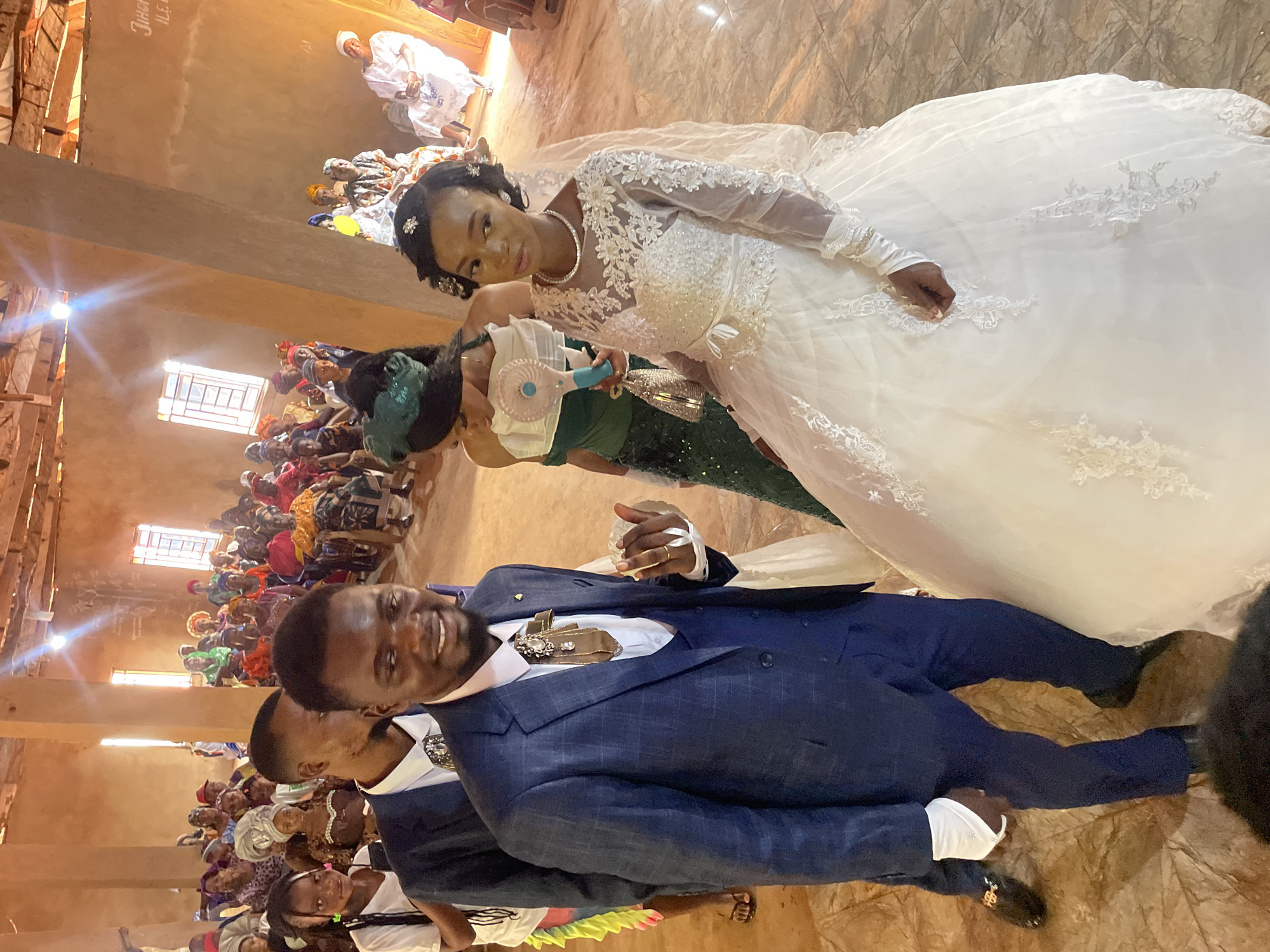 A photograph captures a wedding scene, likely in a church in Ogbe, Nigeria. The bride is adorned in a white gown with lace detailing. She wears a necklace and has hair accessories. The groom is dressed in a blue suit. They are posing for a photo together. In the background, a crowd of people are seated, presumably guests at the wedding. The church's interior features exposed wooden beams and multiple windows, casting natural light.