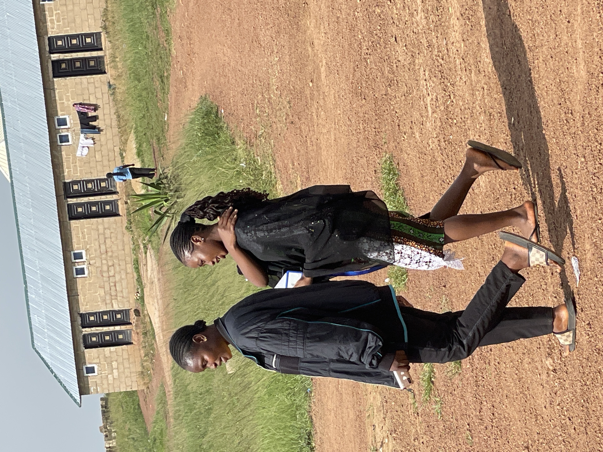 Two young women are walking side-by-side on a dirt path in Durumi, Nigeria. The woman on the left wears a black jacket and black pants. She is holding a white object, possibly a phone or a small book, in her left hand. The woman on the right wears a black, flowing garment over a skirt with a colorful patterned hem. She has her left arm around the shoulder of the woman on the left. Both women have their hair braided.
In the background, there is a low-rise building constructed of light-colored bricks with a corrugated metal roof. The building has several dark-colored doors and small white windows. Laundry is hanging on a line in front of the building. A single person in a blue shirt and dark pants is walking away from the building in the mid-ground. The environment is rural, with tall green grass and sparse vegetation surrounding the dirt path and the building. The sky is overcast with some visible clouds. The lighting suggests it is daytime, likely late afternoon.