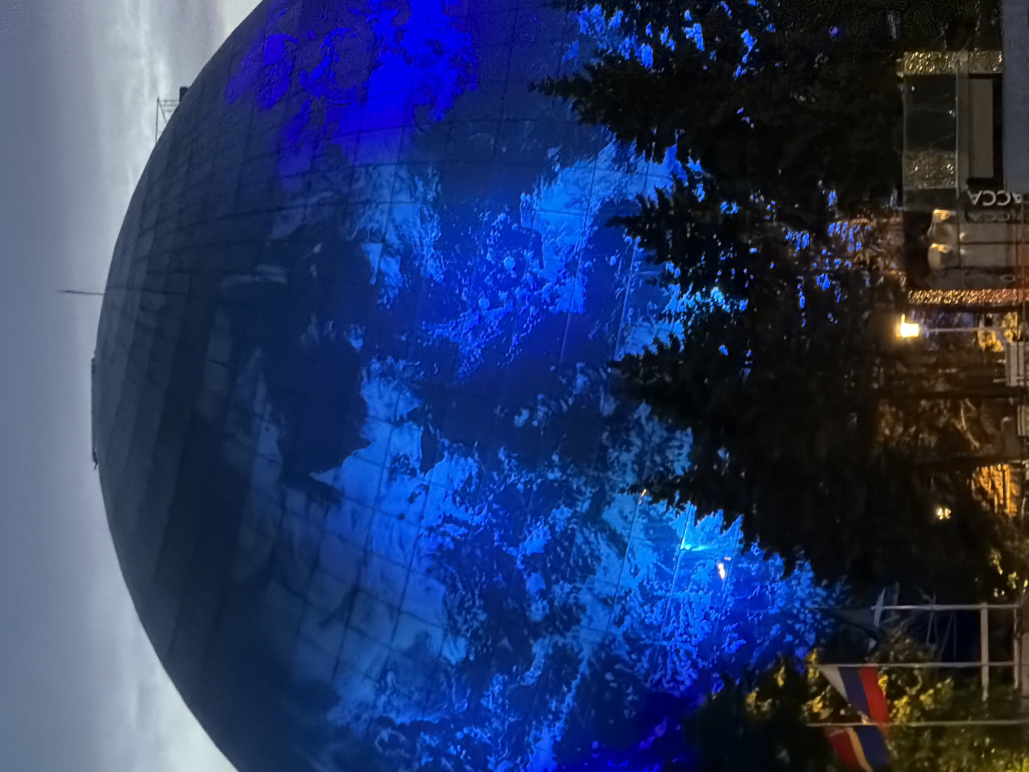 The image shows a large, spherical structure illuminated with vibrant blue light, resembling the Earth. The structure is likely the Kaliningrad Planetarium, which features a large dome. In the foreground, several evergreen trees are silhouetted against the illuminated dome. Two flags, one Russian flag and another with red and yellow stripes, are visible on the left. A lamppost and some illuminated structures, possibly kiosks or stands, are also present in the lower part of the image. The sky is overcast, suggesting it is dusk or night. The scene appears to be an outdoor public space.