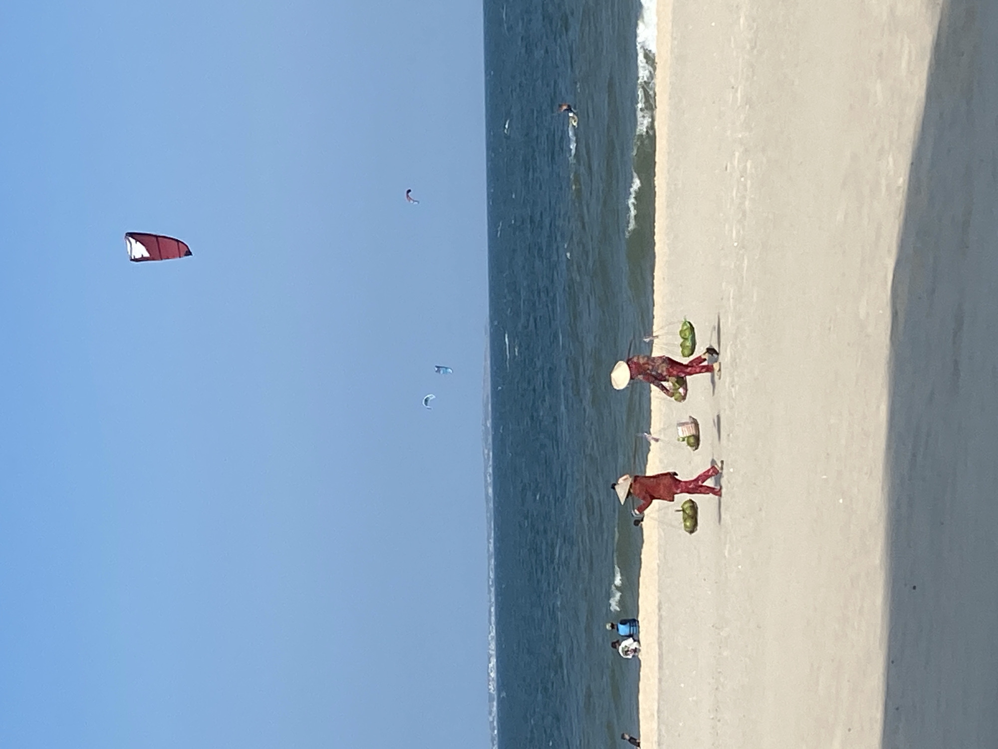 The image displays a sunny day at a beach in Phu Thuy Ward, Vietnam. The sky is clear and bright blue, featuring a large red and white kitesurf kite high in the upper center, and three smaller kites (two blue, one purplish) positioned further right over the ocean. The ocean is a deep blue, exhibiting moderate waves with white caps breaking near the shore. A distant, hazy landmass is visible on the horizon. A single individual is seen lying on a board in the ocean to the right of the image center.
On the light beige sand in the mid-ground, two women are walking from right to left. Both wear conical hats (nón lá) and carry shoulder poles (gánh) from which baskets of green coconuts are suspended. The woman on the left wears a red, long-sleeved outfit, and the woman on the right wears a floral patterned, long-sleeved outfit. Closer to the water's edge on the left, a small group of people are present, including one person wearing a blue life vest and another seated on the sand. A dark shadow partially obscures the bottom edge of the sandy foreground.