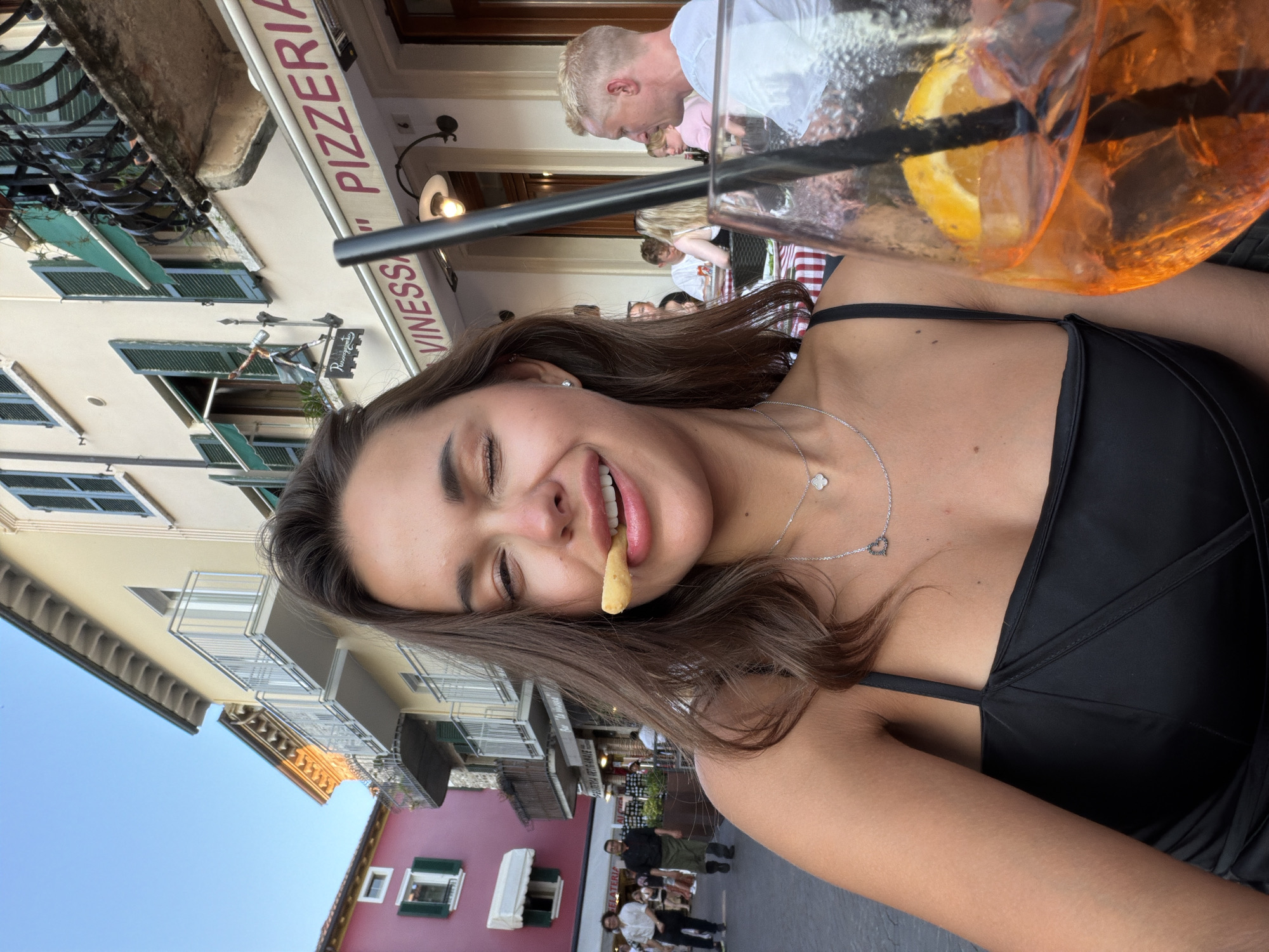 A young woman with brown hair is taking a selfie at an outdoor cafe in Sirmione, Italy. She is wearing a black top and has a piece of food in her mouth, winking at the camera. A tall glass with a bright orange drink and a slice of orange is in the foreground. In the background, there are buildings with balconies and awnings, a sign that reads "Piazzetta", and other patrons dining at tables. The woman appears to be enjoying her meal and the atmosphere of the Italian town.