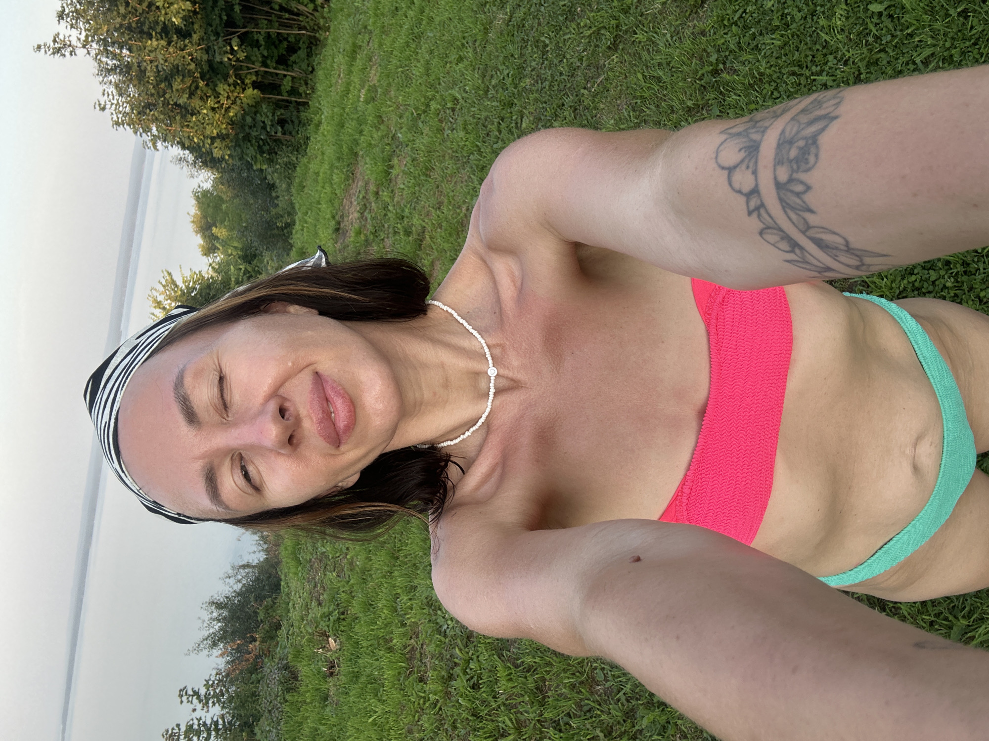 A woman is taking a selfie outdoors. She is wearing a pink bikini top and a teal bikini bottom, along with a black and white striped bandana. A pearl necklace is visible around her neck. She has a tattoo on her upper arm. The background shows green grass and a body of water, likely a lake or river, with trees in the distance. The lighting suggests it is either early morning or late afternoon. The woman is winking and sticking out her tongue, indicating a playful or relaxed mood. The scene is set in Galeyevo, Russia. There is no visible text in the image.