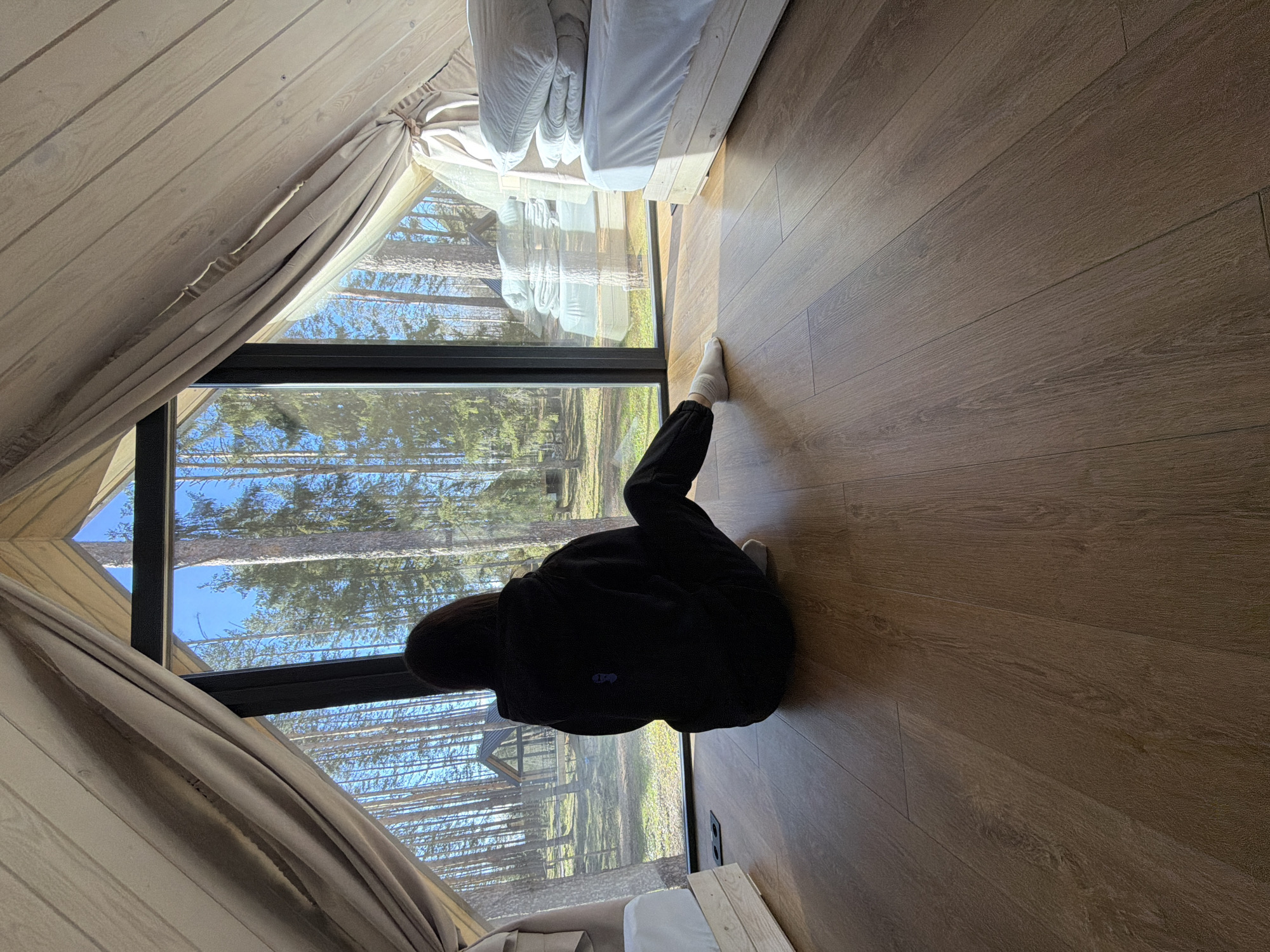 This image captures the interior of a modern cabin or glamping unit in Мишкино, Russia, featuring a large window overlooking a forest. The photograph is taken from a high, tilted angle, looking down into the space.
A person is the primary subject, seen from behind, kneeling or sitting on the light-toned wooden floor. They are dressed in a dark long-sleeved top and dark pants, with white socks visible on their feet. Their posture suggests a moment of calm observation or contemplation as they face the expansive window.
The large window, framed in black, offers a panoramic view of a dense forest composed of tall coniferous trees under a clear blue sky. The bright daylight indicates it is likely morning or afternoon. Sunlight streams into the room, creating subtle shadows on the wooden floor. A reflection in the window reveals more of the forest and hints at another structure, possibly a similar cabin, nestled among the trees.
The interior design is minimalist and natural, with light-colored wooden planks forming the sloped walls and ceiling. On the left side of the window, light beige curtains are neatly tied back, partially framing the view. To the right of the person, a simple bed with white bedding is partially visible. A dark power outlet with two sockets is mounted on the wooden wall near the floor by the window. The overall scene conveys a sense of tranquility and connection with nature. No visible text is present in the image.
