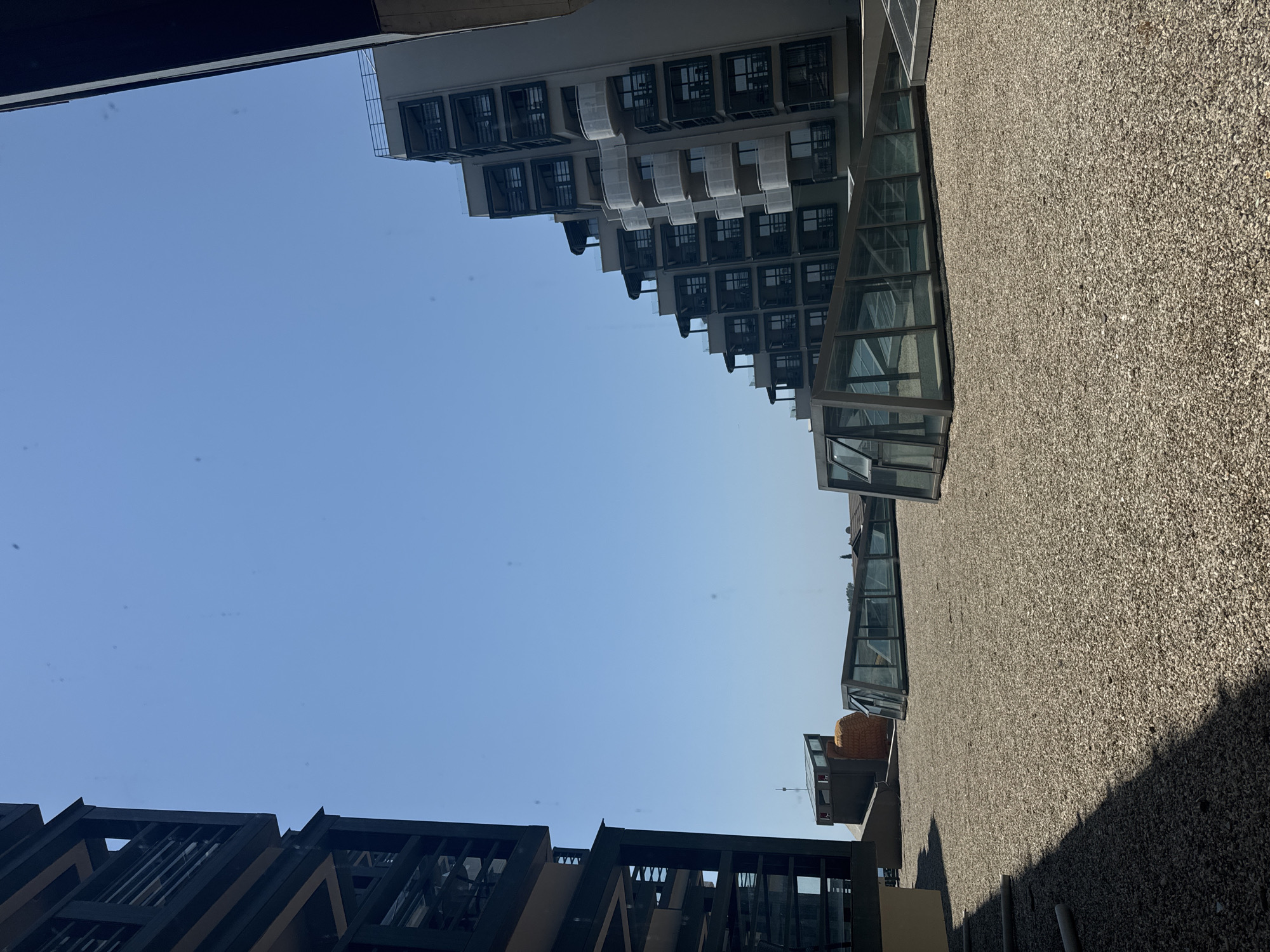 The image captures a rooftop view in Aksu, Turkey, under a clear blue sky. In the foreground, a gravel-covered rooftop is visible, with several angular glass-enclosed structures emerging from it. To the left, a dark, multi-story building with balconies dominates the frame. To the right, a stepped building with numerous balconies, also featuring dark railings, rises against the sky. The architecture suggests a modern urban or resort setting. The scene is devoid of people, focusing solely on the architectural elements and the expansive sky.