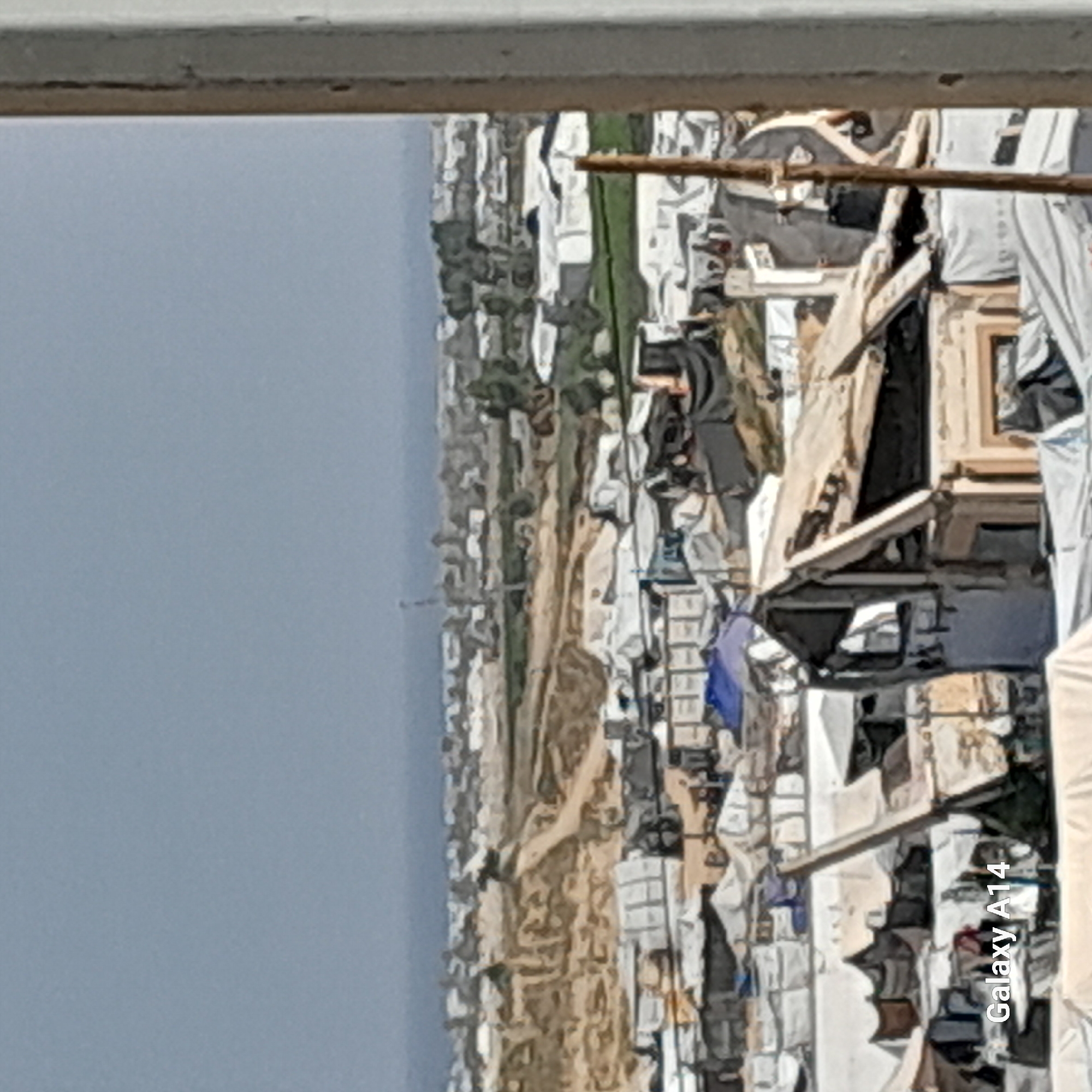 The image, viewed from a high vantage point looking down, depicts a densely packed informal settlement or refugee camp under a clear, bright sky. The scene is dominated by numerous makeshift shelters and structures constructed from various materials, primarily white or light-colored tarpaulins and fabrics, interspersed with darker sections. Some structures appear to incorporate more solid elements like wood or salvaged building materials. A blue tarp or fabric is visible among the shelters in the mid-right area.
Narrow, unpaved paths or strips of sandy, light brown earth wind through the closely arranged dwellings. Beyond the immediate settlement, in the middle distance, there is a stretch of green vegetation, suggesting trees or bushes, followed by a faint outline of a more developed urban area or town under a pale blue sky.
No specific activities or individuals are clearly discernible due to the distance and resolution, though a dark, indistinct figure might be present among the structures in the mid-right. The photograph appears to have been taken during daylight, likely midday, given the bright illumination and lack of long shadows. A railing or ledge is visible across the top of the frame, indicating the photo was taken from an elevated position, possibly a building or platform.
The text "Galaxy A14" is visible vertically aligned along the right edge of the image, indicating the device used to capture the photo.