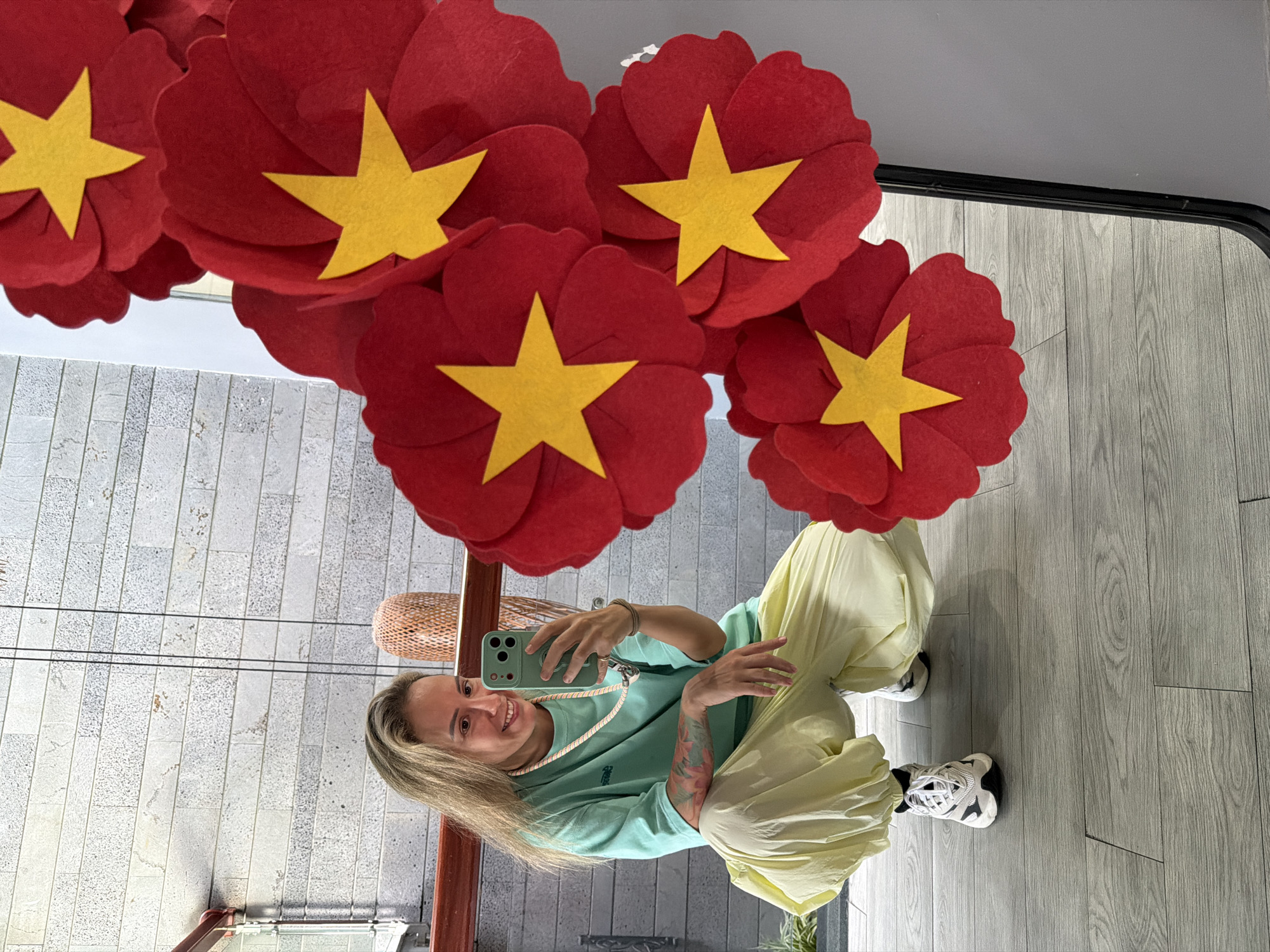 In the city of South Quy Nhon Ward, Vietnam, a young woman with blonde hair is crouched down, taking a selfie in a mirror. She is wearing a light green t-shirt, loose pale yellow pants, and white sneakers. A beaded necklace is around her neck. The background shows a textured grey wall and a wooden floor. In the foreground, a large decorative piece made of red felt flowers with yellow stars is visible, partially obscuring the reflection in the mirror.