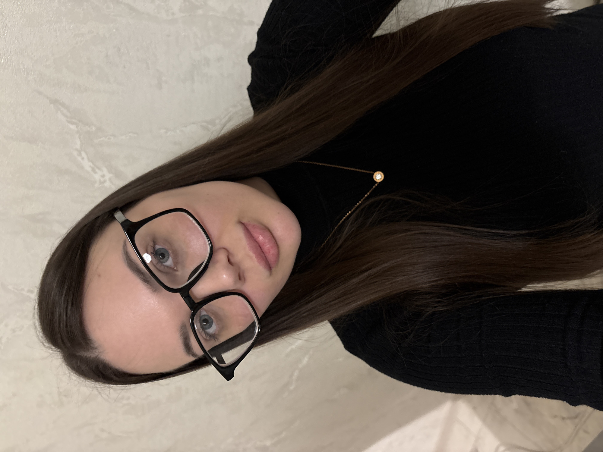 A person is centered in an indoor close-up shot. They wear a black turtleneck sweater and glasses with a black frame. Their hair is long, straight, and brown, and they have blue eyes. A gold necklace with a single circular pendant is visible around their neck. The background appears to be a light-colored wall. The image is oriented at a slight angle. There is no definitive information in the image that indicates the location is Chaikovsky, Russia.