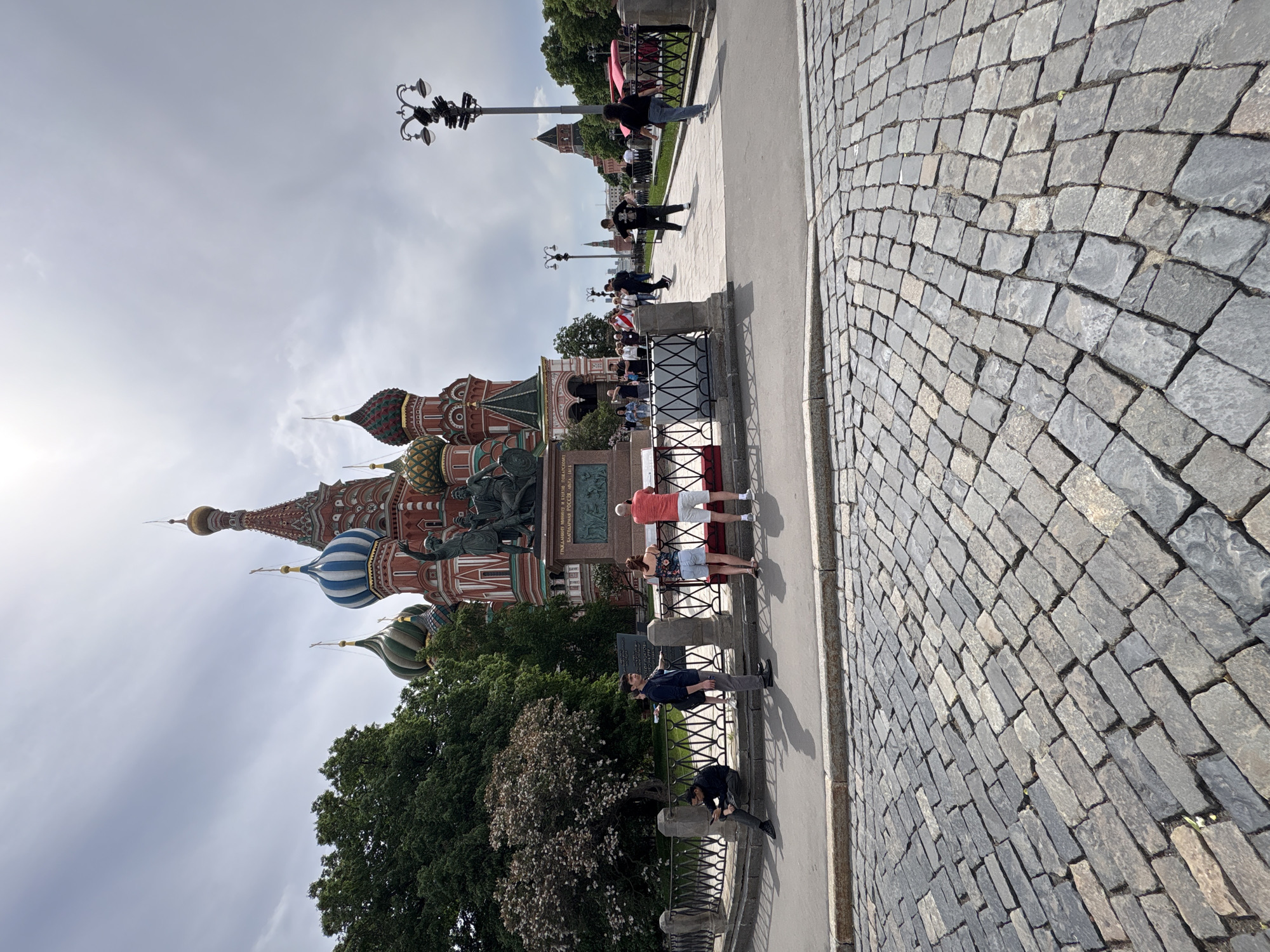 The image depicts a street scene in front of St. Basil's Cathedral in Moscow, Russia. The foreground features a cobblestone street with a few people walking and standing nearby. The cathedral itself is visible in the background, showcasing its colorful domes and architecture. Some trees and a lamppost are also visible, adding to the overall scene.