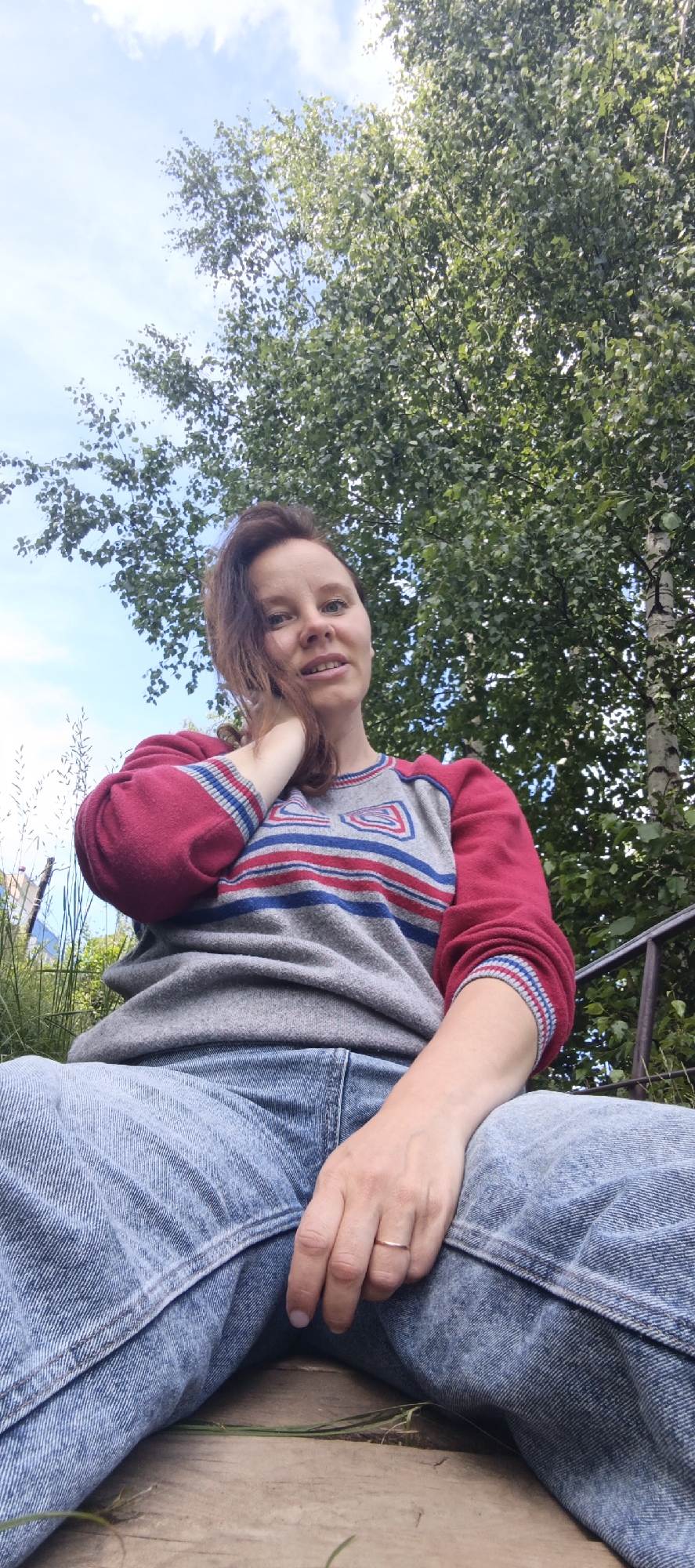 A woman with auburn hair sits outdoors in a low-angle shot, looking towards the camera. She is wearing a grey sweatshirt with red sleeves and blue and red stripes on the chest, and light blue denim jeans. Her right hand is raised and touching her neck, and she is wearing a thin, rose-gold colored ring on her left ring finger. The background consists of green trees and a bright blue sky with scattered white clouds, suggesting a sunny day. A glimpse of what appears to be a metal railing is visible to the right of the woman. The perspective suggests she is sitting on a wooden surface, possibly a deck or stairs. The scene conveys a relaxed and casual mood.