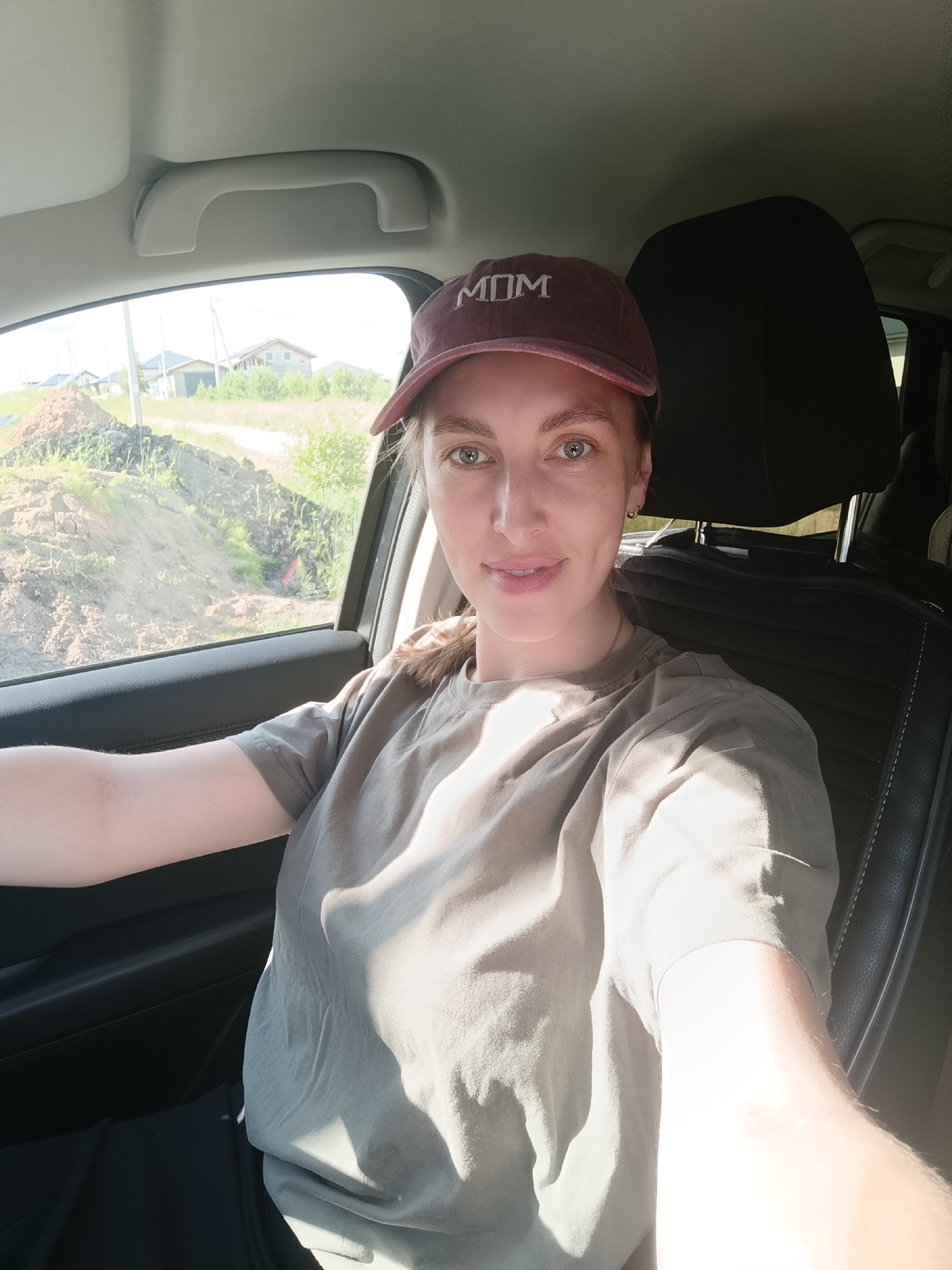 This is a waist-up, front-facing selfie of a young woman sitting in the driver's seat of a car. She is wearing a maroon baseball cap with the word "MOM" embroidered in white on the front. She also has on a light brown t-shirt. Her left arm is resting on the steering wheel, and her right arm is extended towards the camera. She has light skin, blue eyes, and light brown hair that is pulled back and visible under her hat and over her shoulders. She is smiling slightly.
The background shows the interior of the car, with the car seat behind her. Through the window on her left, a rural landscape is visible. There are some unfinished houses, construction debris, and green vegetation. The scene suggests it is daytime, likely a sunny day due to the strong shadows and bright lighting on the woman's face and t-shirt. The location context of Chaikovsky, Russia, places this in a potentially rural or suburban area. The woman appears relaxed and cheerful.