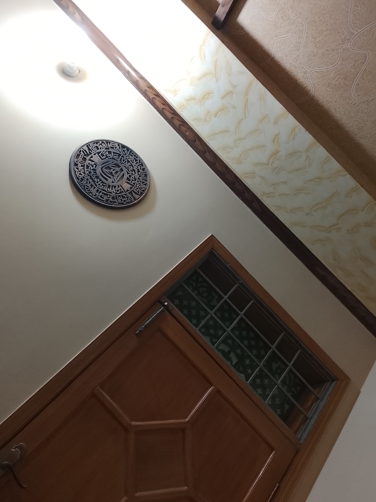 A low-angle shot depicts a door and adjacent wall. The door is wooden, with a window pane section above and a panelled lower section. The door is closed. A metal latch mechanism is visible on the upper left of the door. To the left of the door and near the top of the frame, a decorative round plaque hangs on the wall, featuring Arabic script. Above the plaque is a ceiling light fixture. A dark wooden beam runs diagonally across the upper portion of the frame, adjacent to a patterned wall section. The photograph was taken indoors. The location is identified as Multan, Pakistan.