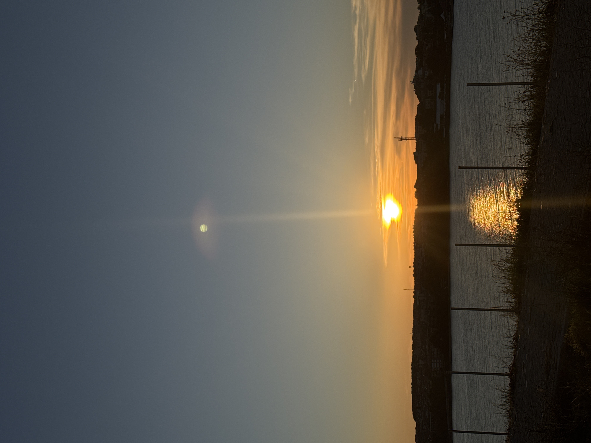 A vertical, low-angle shot captures a serene sunset over a body of water, likely the sea, in Tsarevo, Bulgaria. The sky transitions from a deep, twilight blue at the top to a warm, gradient of orange and yellow near the horizon. A bright, orb-like sun is partially visible, casting a shimmering golden reflection across the water's surface. The shoreline is silhouetted against the vibrant sky, revealing a cluster of buildings and a prominent cell tower on a gentle incline. In the foreground, the dark, textured ground is dotted with dry grass and is intersected by a line of simple, vertical posts. There are no people or animals present in the image. The overall mood is one of peaceful contemplation, highlighting the natural beauty of the coastal landscape during the golden hour.