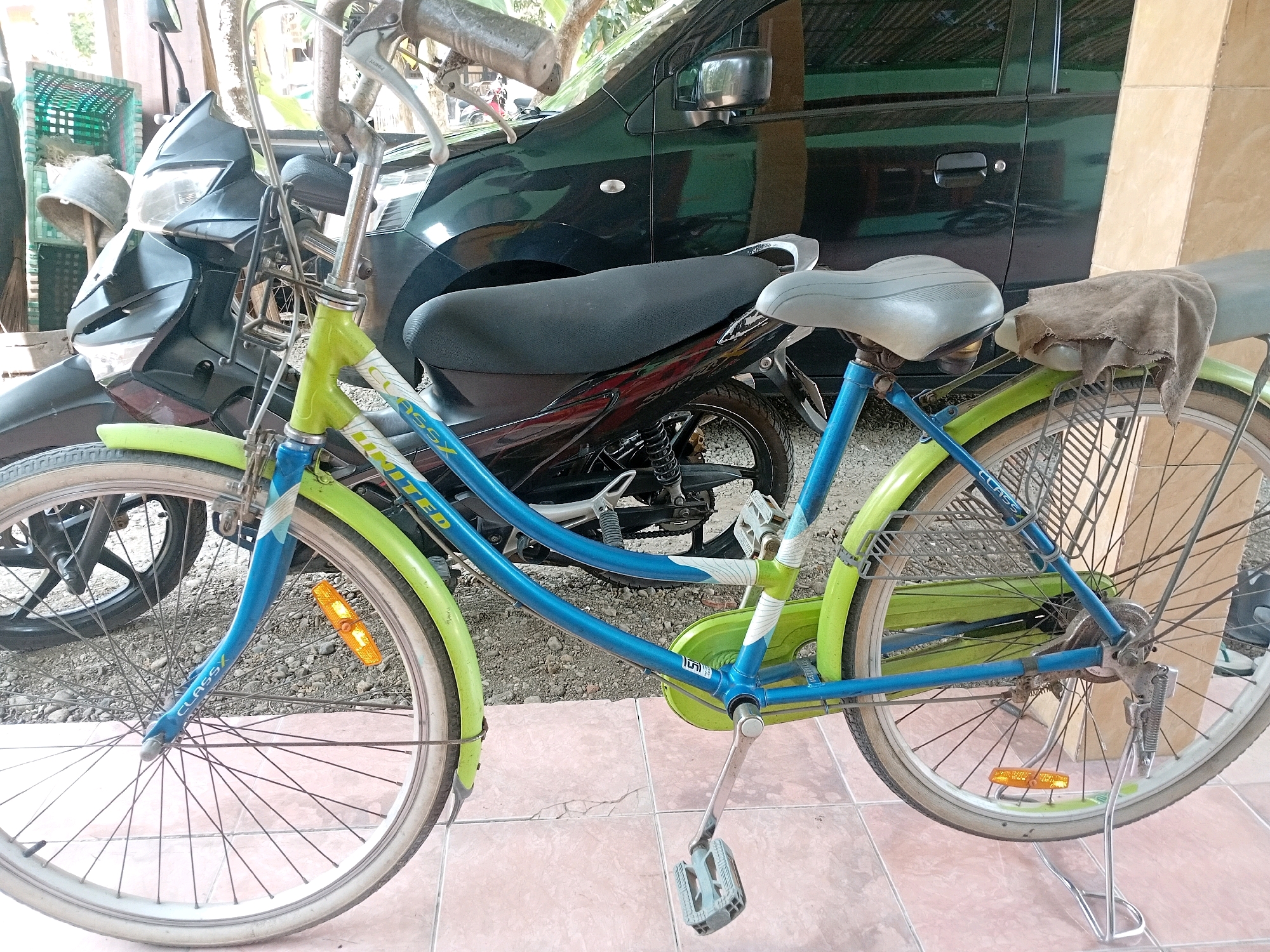 The image shows a blue and green bicycle parked next to a black motorcycle and a black car, likely on a residential property in Aditirto, Indonesia. The bicycle has a basket on the rear, a grey seat with a rag draped over it, and reflective orange markers on the front and rear wheels. The motorcycle is positioned behind the bicycle, with its front end visible. The car is parked to the right, partially obscuring the view of the motorcycle. The ground is tiled in some areas and gravel in others. In the background, there are crates and foliage, suggesting an outdoor setting.