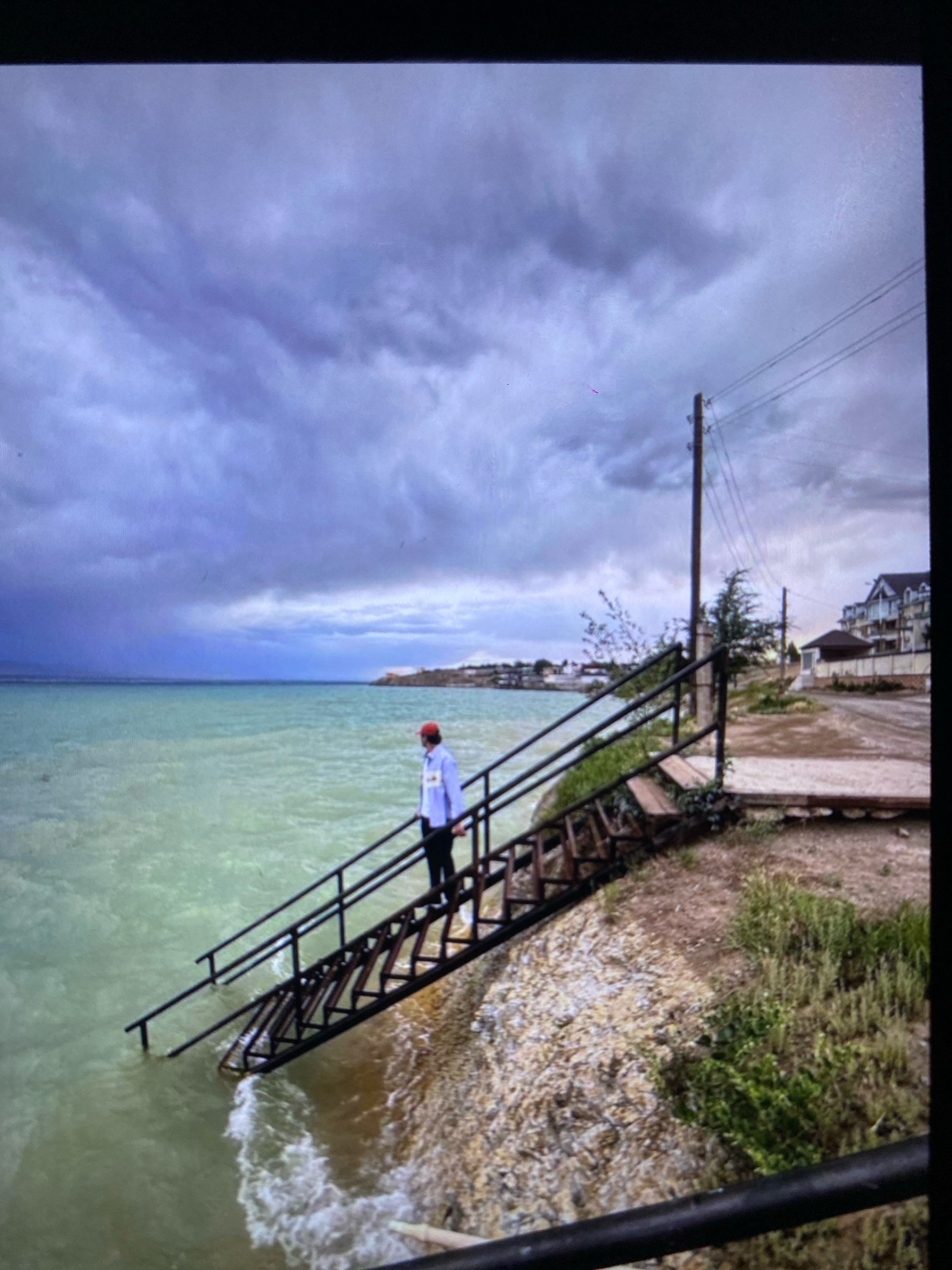 A person stands on a metal staircase that descends into the turquoise water of a lake. The staircase is situated on a gravelly shore dotted with dry grass and a few small bushes. The sky above is filled with dramatic, dark grey clouds, suggesting an approaching storm or overcast weather. In the background, a few buildings can be seen along the distant shoreline, and a utility pole with overhead wires stands to the right of the staircase. The water appears calm, with gentle waves lapping at the base of the stairs. The scene is devoid of any specific landmarks that would place it within the city of Ramenskoye, Russia.
