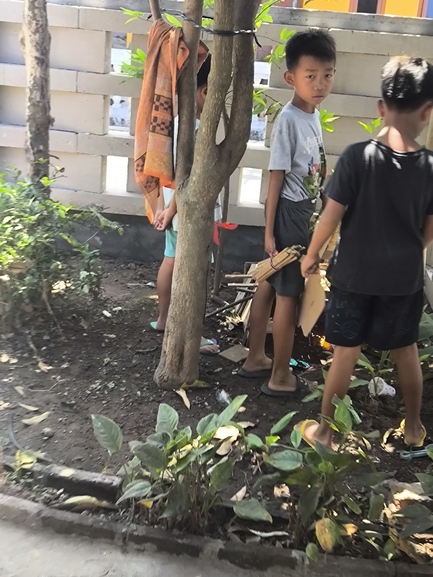 In Kebonsari, Indonesia, a group of children stand in a dirt yard with scattered debris and sparse vegetation. Two young boys are the primary subjects. The boy on the left, facing forward, wears a grey t-shirt and dark shorts, holding a bundle of thin wooden sticks. The boy on the right, viewed from behind, wears a black t-shirt and dark shorts. Another child, partially obscured by a tree, wears a light blue garment and teal shorts, with an orange towel draped over their shoulder. A rough brick wall forms the background, with a tree trunk in the foreground. The ground is a mixture of dirt, leaves, and various small objects.