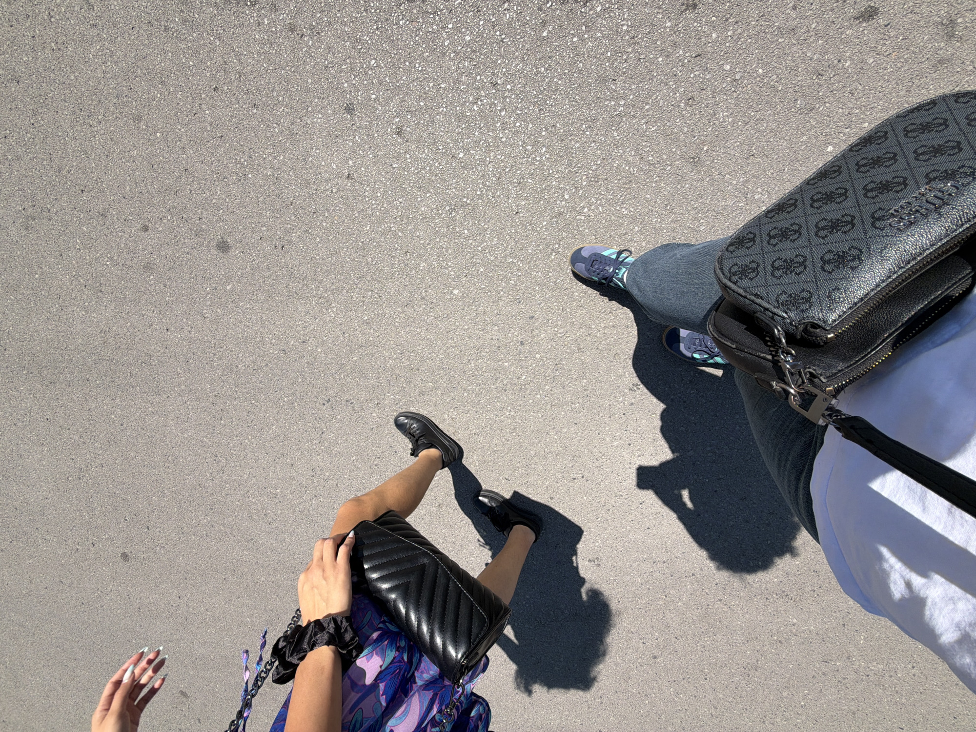 Two individuals are walking on an asphalt surface, likely a street or sidewalk, in Sofia, Bulgaria. The first individual, on the left, is wearing a vibrant purple and blue patterned dress and black shoes. They are carrying a quilted black purse in their right hand and have long, manicured fingernails visible. A black scrunchie is also visible on their wrist. The second individual, on the right, is wearing dark denim jeans and athletic shoes with blue and green accents. They are carrying a black crossbody bag with a repeating Guess logo pattern. Both individuals are casting shadows on the ground, indicating a sunny day. The environment is predominantly the grey asphalt surface, with some subtle variations in texture and color.