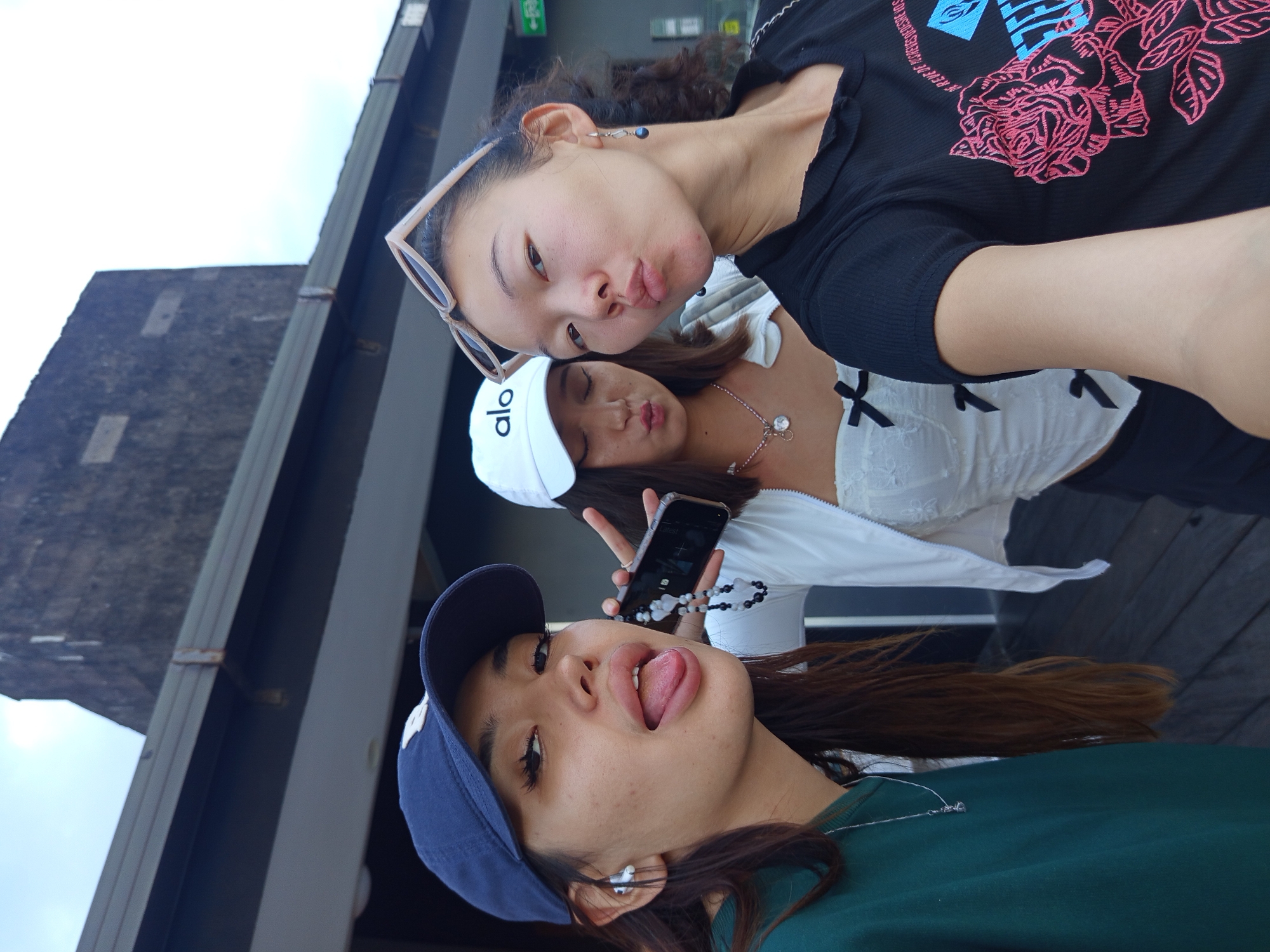 Three young women are taking a selfie in North Kuta, Indonesia. The woman closest to the camera wears a navy blue baseball cap, a green shirt, and silver earrings. She has her tongue out and is holding a cell phone with a beaded wrist strap. Behind her, a woman in a white “Alo” baseball cap and white top is making a kissy face. The third woman, at the top of the frame, also has her lips pursed and wears sunglasses on her head and a black top with a pink rose graphic. In the background is a modern, dark-colored building structure.