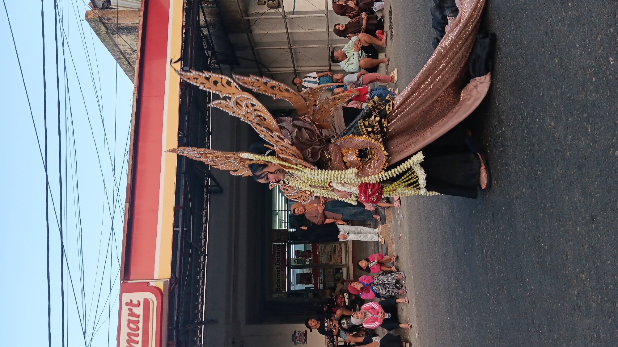 In Tanjung Anom, Indonesia, a parade features a young woman in an elaborate, winged costume. The costume, adorned with gold and brown sequins, white flower garlands, and red roses, accentuates her stature. She is positioned on a street, likely during a local festival or event. Spectators, including children and adults, are seated on the curb and stand near a brightly colored storefront, identified by a "mart" sign. The background reveals buildings and overhead electrical wires, characteristic of a bustling town. The overall scene captures a vibrant cultural celebration, with the central figure commanding attention amidst the onlookers.