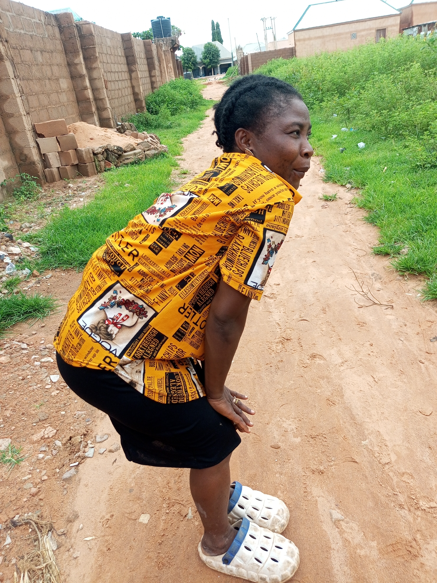 An outdoor, eye-level, medium shot captures a Black woman standing on a dirt path in what appears to be a rural or developing area of Tempure, Nigeria. The woman is facing right, with her body angled slightly away from the camera. She is wearing a bright yellow shirt with a busy pattern of text and images, a black skirt, and white Croc-like shoes with blue accents. Her hair is styled in dark braids. She has her hands on her knees, and her head is turned to look back over her shoulder.
The environment is characterized by dry, sandy soil forming a path that curves to the right. On the left side of the path, there is a partially constructed brick wall and a pile of sand and bricks, suggesting ongoing construction. Beyond the wall, there are some patches of green grass and dry weeds. On the right side of the path, dense green vegetation grows, with some litter visible. In the background, there are buildings with light-colored walls and roofs, and trees. The sky is overcast. The overall impression is of a functional, possibly residential, area undergoing development.
