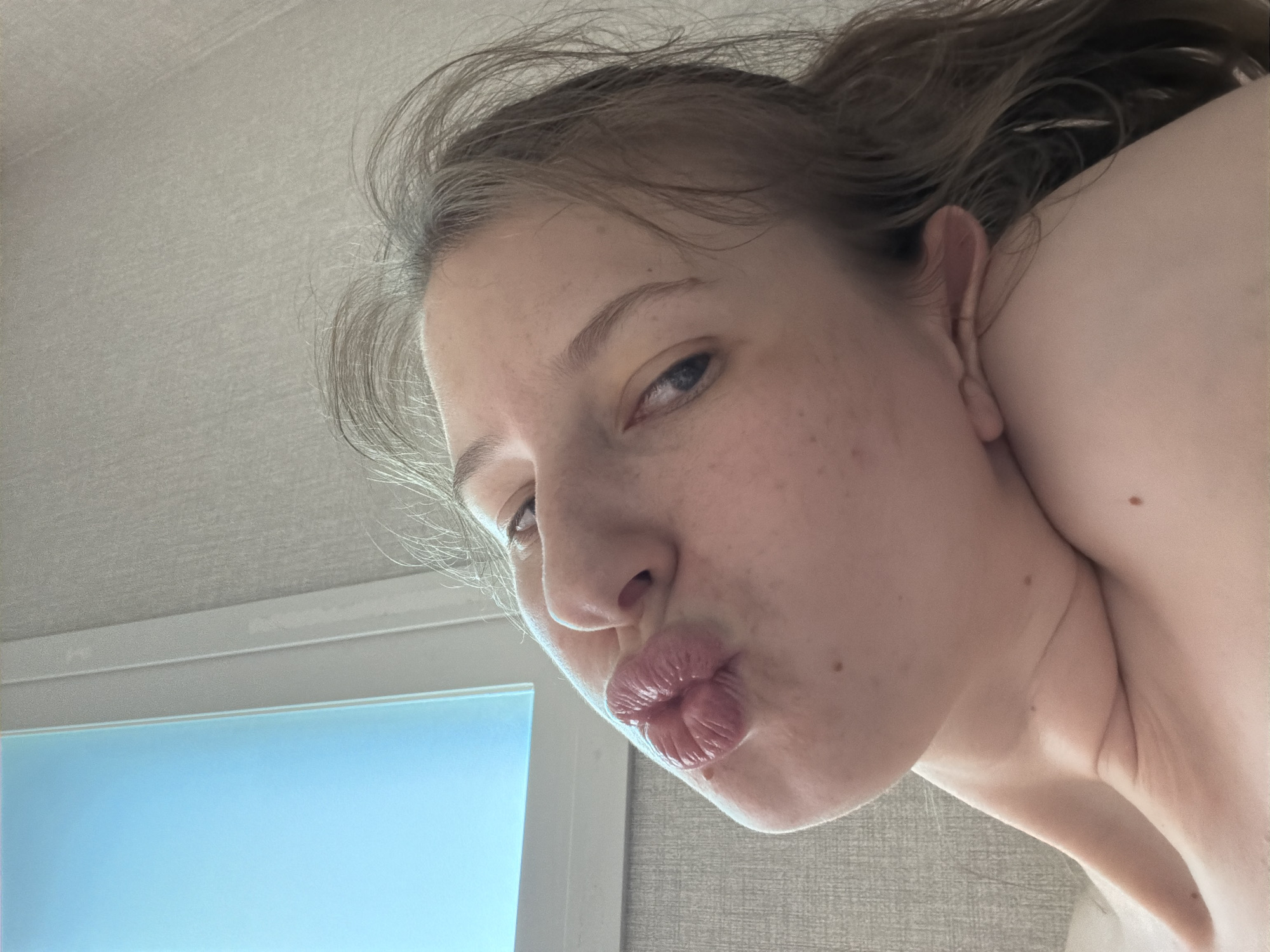 The image shows a close-up, low-angle selfie of a young person with their lips puckered in a kissing gesture. The background is a textured, neutral-colored wall with a bright blue window frame on the left. The person's face is tilted upwards and to the right, with their hair falling loosely around their head and shoulders. Some light freckles are visible on their skin. The overall lighting is soft and diffused. There is no information in the image to suggest it was taken in Dongducheon-si, South Korea.