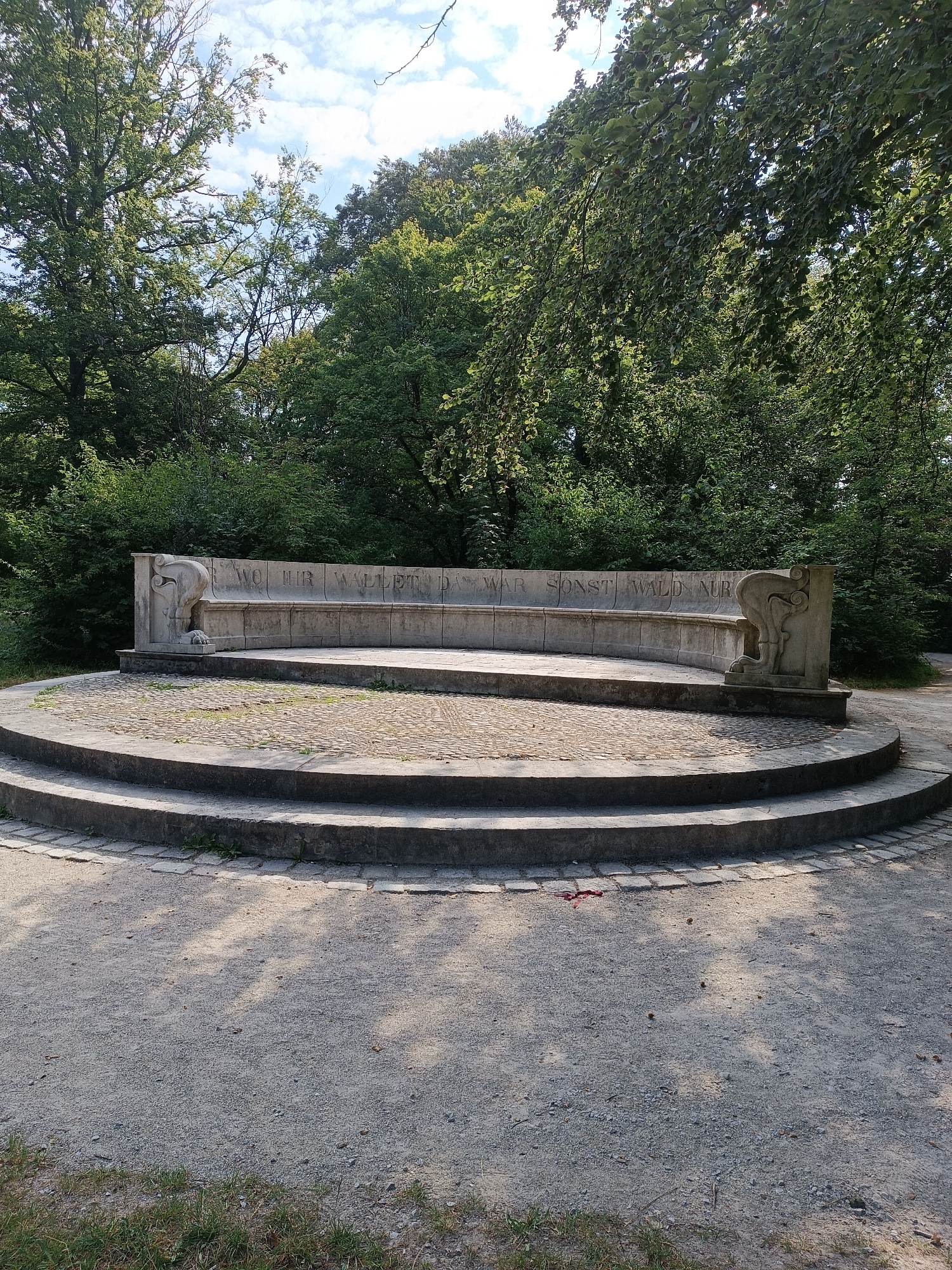 The image shows a semi-circular stone bench with inscribed text, situated in a park-like setting. The bench is elevated on a tiered, circular stone platform with steps leading up to it. The bench itself is carved with decorative elements on either end, resembling stylized animal heads. The inscribed text on the bench reads "WO HIER WALDT DA WAR SONST WALD NUR". This monument is located in the Olympiapark in Munich, Germany, specifically in the area of the Olympiaberg, the artificial hill created from rubble after World War II. The background consists of lush green trees, suggesting a sunny day with some scattered clouds visible in the sky. The ground around the platform is covered with gravel and sparse grass.