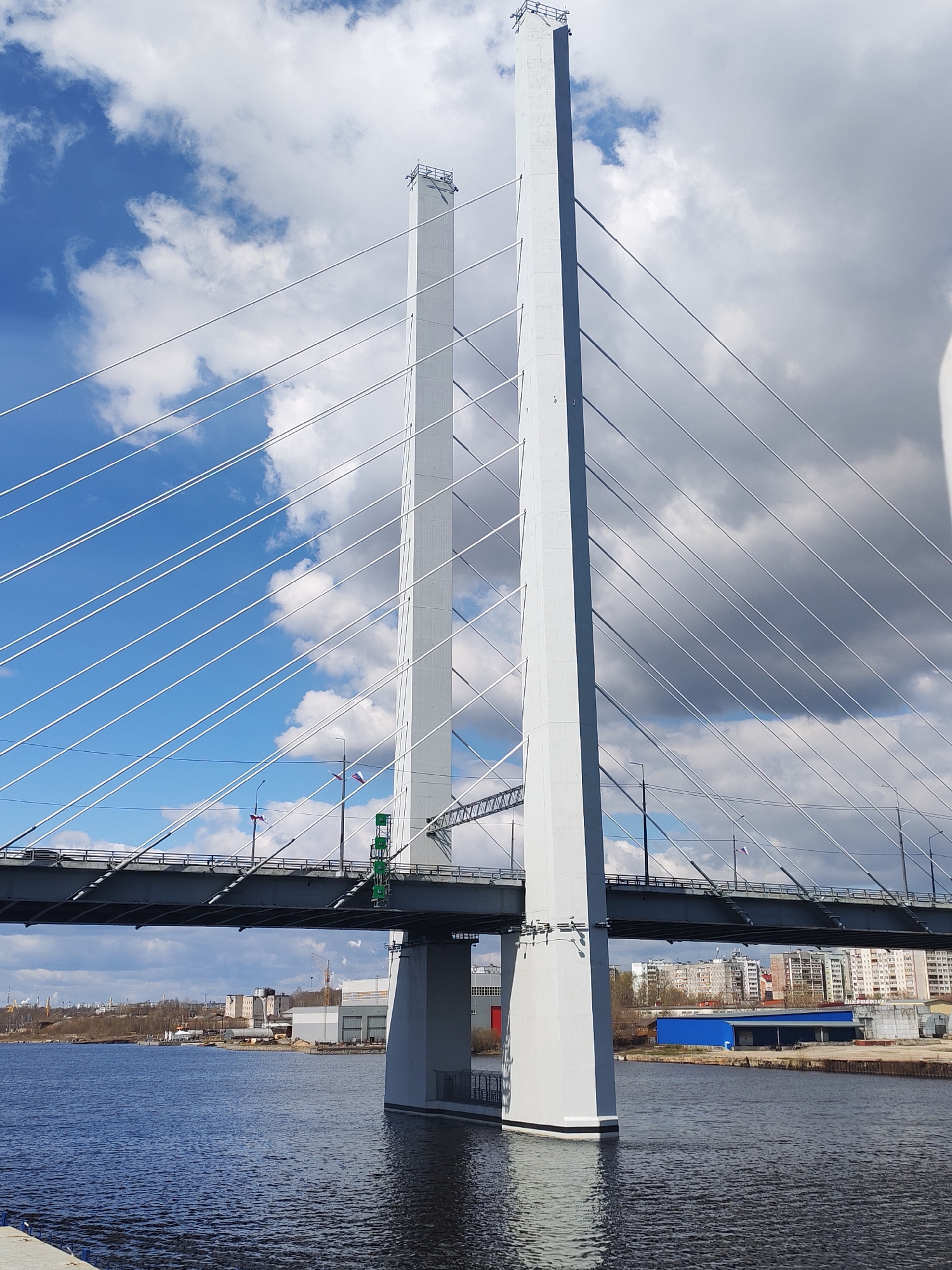 The image depicts a cable-stayed bridge in Cherepovets, Russia, spanning a body of water. Two tall, light-gray concrete pylons rise from the water to support the bridge deck with numerous white steel cables. The bridge deck is a darker gray. Buildings are visible in the background on both sides of the water, along with bare trees suggesting early spring. The sky is a mix of blue and white clouds. The water is dark with subtle ripples. Small Russian flags are attached to light posts on the bridge.