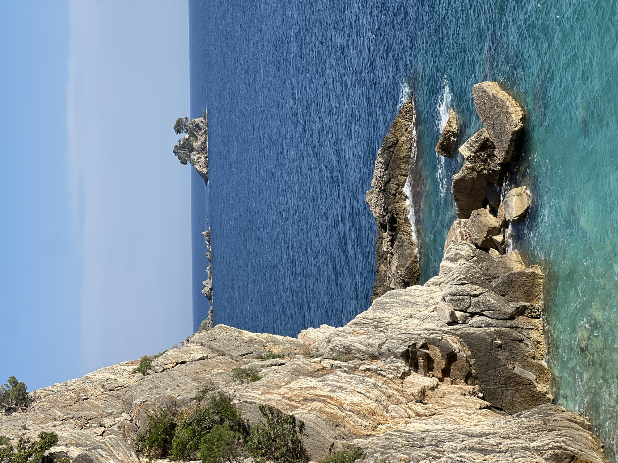 This image depicts a rocky coastline in Perazića Do, Montenegro, with a view of the Adriatic Sea. The foreground shows layered rock formations with sparse vegetation. The sea transitions from deep blue to turquoise near the shore, where rocky outcrops meet the water. In the distance, there is a small, tree-covered islet. The sky is a pale blue with a slight gradient. No people are visible.