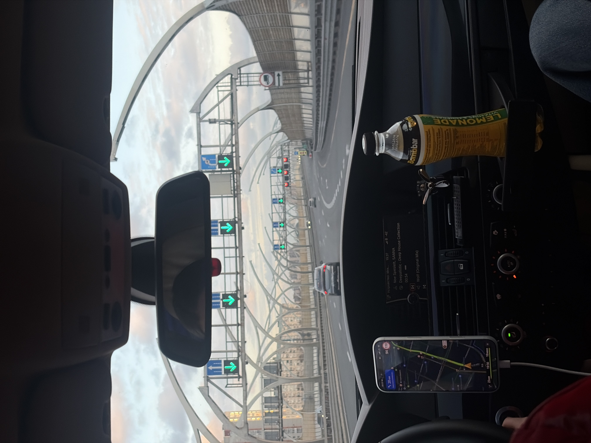 Captured from the interior of a vehicle, the image presents a forward-facing view of the Western High-Speed Diameter (WHSD), a toll highway in Saint Petersburg, Russia. The driver's perspective encompasses the road ahead, lined with distinctive arching structures supporting traffic signage and lighting. Digital displays overhead exhibit green arrow indicators, directing traffic flow. In the foreground, the car's dashboard is visible, featuring a smartphone mounted with a navigation app displaying a route. A bottle of "Bombbar Lemonade" is situated near the car's console. The image suggests the vehicle is in motion, proceeding along the WHSD, a notable infrastructure component of Saint Petersburg's transportation network.
