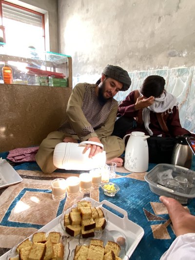 The image depicts an indoor gathering, likely a social occasion, featuring two men and various refreshments. In the foreground, a man with a beard and wearing a dark turban, a light brown tunic, and a patterned vest, sits on a blue and brown geometric-patterned rug. He is holding a white thermos, from which he appears to be pouring or serving a beverage. Several small glasses containing a light-colored drink (possibly tea with milk) are lined up beside him. A small glass bowl filled with yellow items, possibly candies or nuts, is also present.
Two white trays heavily laden with square slices of cake, resembling pound cake, occupy the immediate foreground. Another man is visible in the background, dressed in a dark red garment with a light-colored scarf around his head or neck, his face partially obscured. A third person's foot, wearing white, is visible in the bottom right corner. Other items on the rug include a white thermos with a floral design and a metallic/black thermos, as well as a grey plastic basket containing more glasses.
The setting is a simple room with unpainted or concrete walls. Bright sunlight streams through a window on the left side of the frame, suggesting it is daytime. Below the window, there is a low, light brown counter or shelf topped with a glass display case containing various bottles and other items. The scene suggests a relaxed and informal gathering focused on hospitality and sharing refreshments. No visible text is discernible in the image.