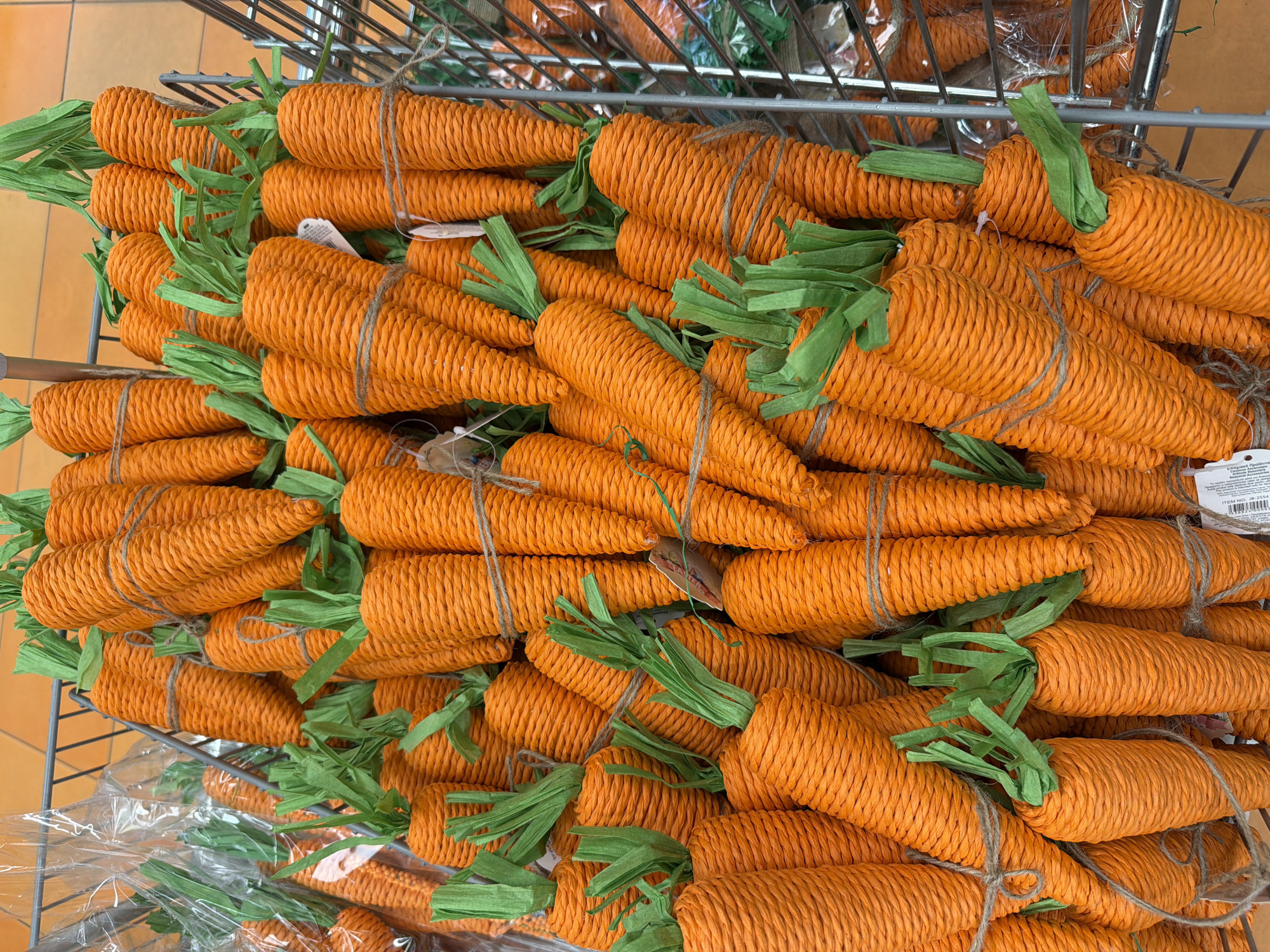 A high-angle, indoor shot shows a densely packed wire shopping cart filled with numerous artificial carrots. The carrots are bright orange, shaped like cones, and textured with tightly wound material. Each has a green, paper-like top resembling carrot leaves, and some are tied with a piece of twine. The cart's wire frame is visible in the background, along with more carrots stacked underneath, some still wrapped in clear plastic. The lighting is bright and even. There is no indication in the image that the location is in the city of Sofia, Bulgaria.