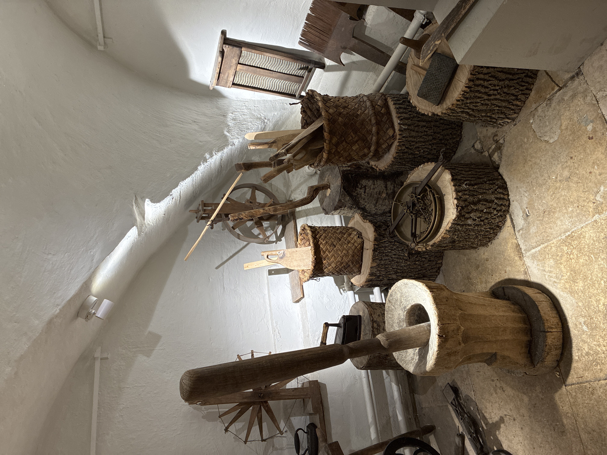 The image depicts a collection of antique wooden tools and implements displayed along a white wall and a stone floor, likely in a museum or historical exhibit. The tools are varied, including what appears to be a washboard, a wooden basket filled with various wooden tools, a wooden wheel, and a large mortar and pestle. The lighting is uneven, creating areas of both bright illumination and deep shadow. The location in Moscow, Russia, is not evident from the image, unless the particular items are distinctly Russian.