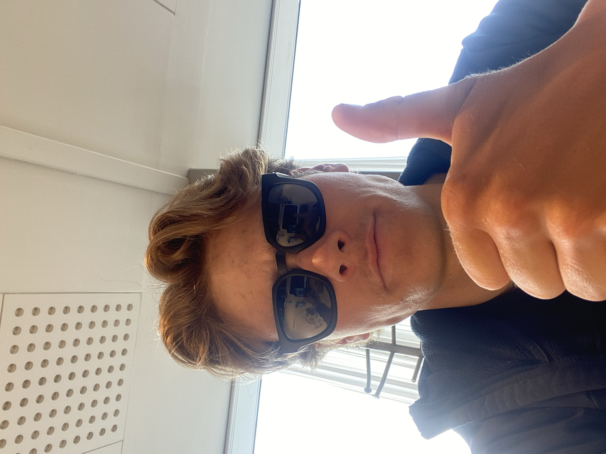 A person with light skin and brown hair is wearing sunglasses and giving a thumbs-up. They are indoors, next to a window. To the left is a white wall with circular perforations. The location is Kastrup, Denmark.