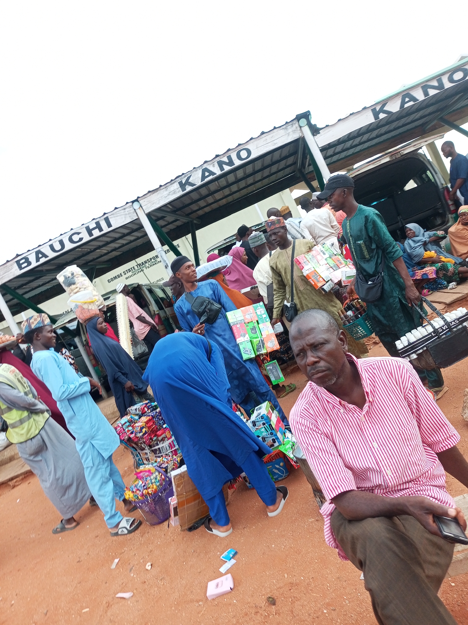 The image depicts a bustling market scene in Gombe, Nigeria, likely at a transportation terminal. A sign above reads "BAUCHI KANO," indicating different destinations or areas served by the terminal. People are engaged in various activities, including selling and buying goods. Several individuals are seen wearing traditional clothing, such as long robes and head coverings. Some are carrying items on their heads or in their hands. The ground is sandy and reddish-brown, typical of the region. Stalls are set up with a variety of merchandise, including packaged goods, baskets filled with items, and what appear to be electronics or small accessories. The atmosphere suggests a busy marketplace with a sense of movement and commerce. A van is visible in the background, suggesting transportation services are available. The overall scene is lively and indicative of daily life in a Nigerian urban center.