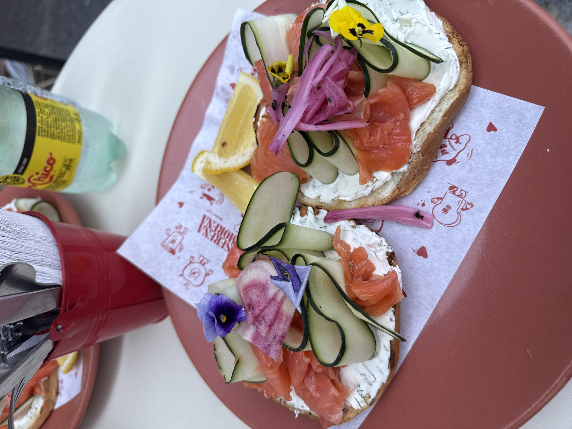 The image shows two open-faced sandwiches on a terracotta-colored plate. Each sandwich consists of a toasted base spread with white cream cheese, topped with slices of smoked salmon, cucumber ribbons, and colorful edible flowers. One sandwich also has pickled red onions and lemon slices. The sandwiches rest on a square piece of white paper printed with red cartoon-like characters and the words "SERENA MORENA." A partially visible green glass bottle with a yellow label is on the left side of the frame. Next to it is a small red bucket containing metal cutlery and napkins. The scene is likely in Puebla, Mexico, given the name "SERENA MORENA" printed on the paper.