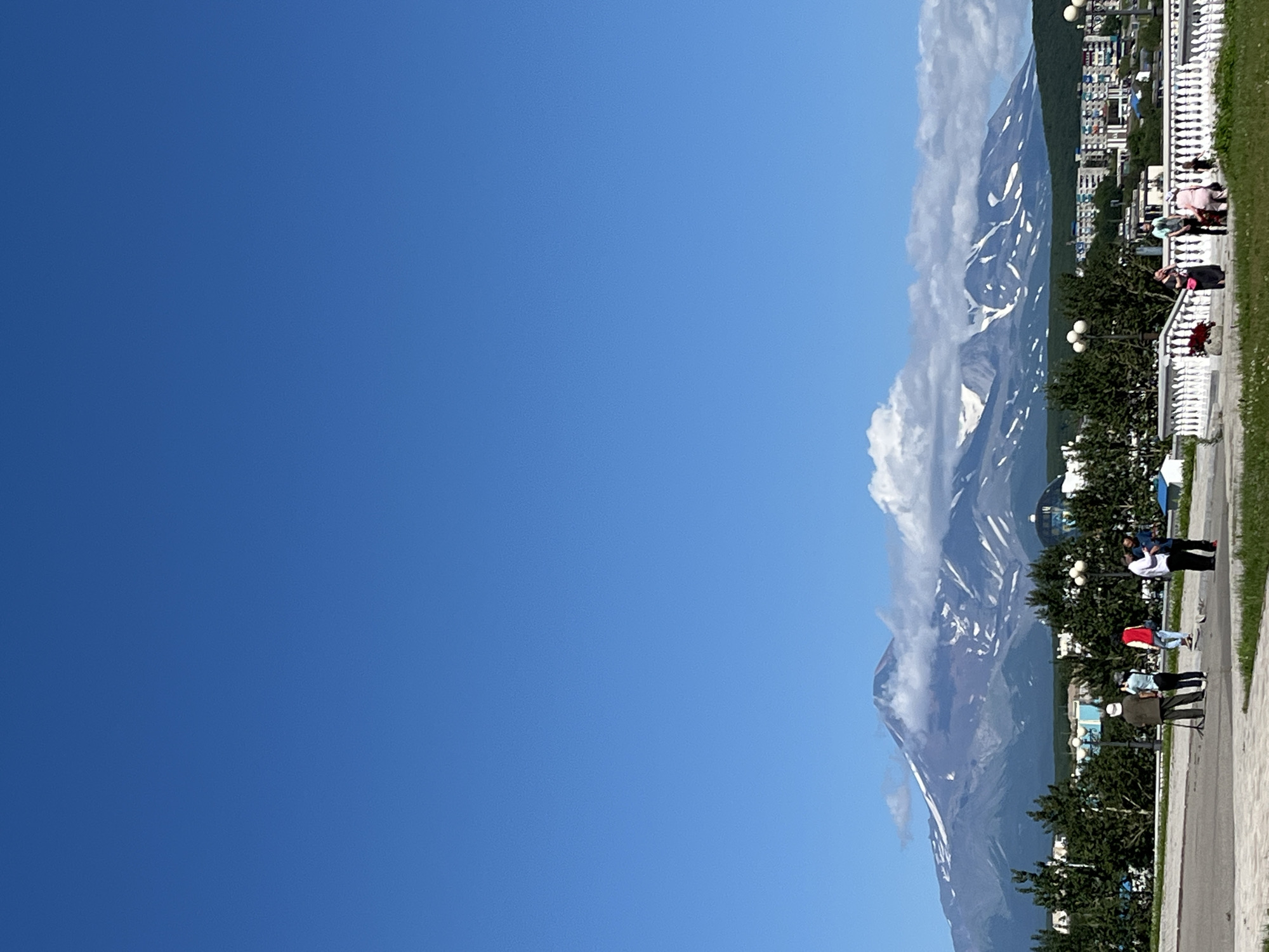 The image shows a bright, sunny day with a clear blue sky. In the background, a majestic mountain range, likely volcanoes, is visible, with some snow patches on their peaks and white clouds partially obscuring them. In the foreground, a street or promenade area is depicted, with a white railing and lampposts. Several people are scattered in the foreground, some standing and observing the scenery, while others appear to be walking or conversing. There are trees and buildings in the middle ground, suggesting an urban or semi-urban setting. The overall impression is of a scenic vista in the city of Petropavlovsk-Kamchatsky, Russia, known for its proximity to active volcanoes.