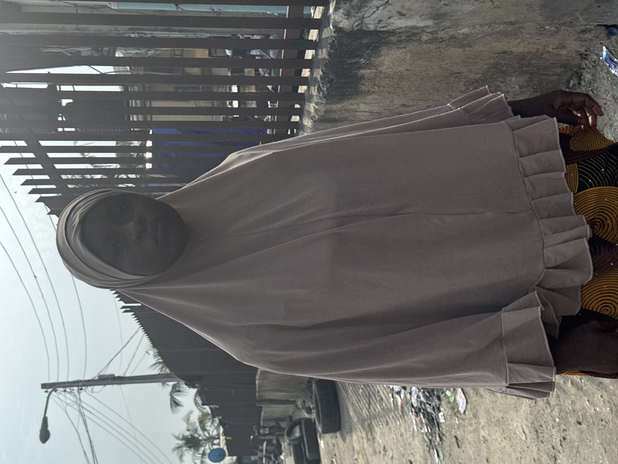 The image, rotated 90 degrees counter-clockwise, depicts an individual, likely a young woman, standing outdoors in what appears to be an urban or semi-urban environment in Ado, Nigeria.
The subject is facing the camera, wearing a light beige or light brown hijab that covers her head and drapes over her upper body. The garment has distinctive ruffled cuffs or edges on the sleeves. Beneath the hijab, a dress or skirt made of fabric with a vibrant brown and yellow circular or spiral pattern is visible. Her expression is neutral as she looks directly at the viewer.
The setting includes a rough concrete ground with scattered debris or litter. In the background, a dark metal fence or gate with horizontal slats is prominent, partially obscuring the area behind it. To the left of the subject (when the image is correctly oriented), a utility pole with multiple overhead wires stands against a bright, hazy sky, indicating a daytime scene. The lighting suggests bright sunlight, possibly with some haze. A dark car tire is also partially visible on the ground.
No specific activity is taking place other than the individual posing for the photograph. There is no visible text in the image.