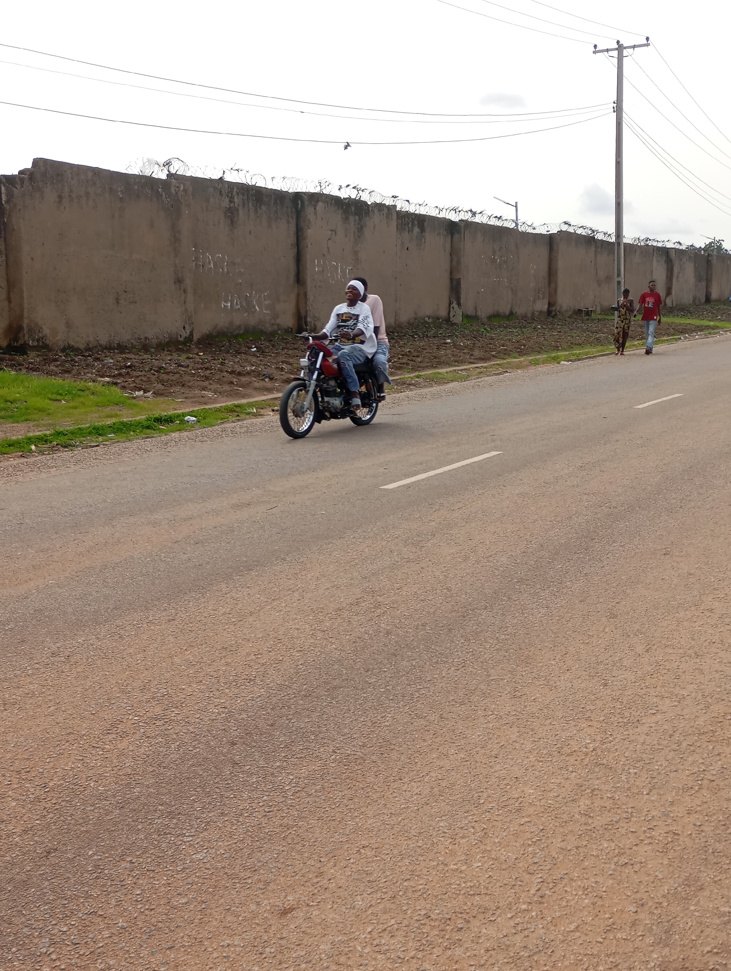 An eye-level, medium shot captures a street scene in Shendam, Nigeria, under an overcast sky. In the foreground, a light-colored asphalt road stretches across the frame, marked with a single white lane divider. To the left, a patch of green grass and dirt lies between the road and a large, rough concrete wall topped with barbed wire. The wall has graffiti in what appears to be white paint, with words like "HACKE" visible.
A red motorcycle with two occupants rides along the road, moving from left to right. The person in the front, wearing a white long-sleeved shirt with a graphic design and blue jeans, is driving. The passenger behind them is wearing a white shirt and blue jeans, with a white headband. They are both looking ahead and appear to be smiling.
Further down the road, a woman in a patterned dress and a man in a red t-shirt and blue jeans are walking away from the motorcycle, also moving from left to right. A utility pole with power lines stands on the right side of the road, behind the pedestrians. The overall environment suggests a somewhat dry, dusty area.