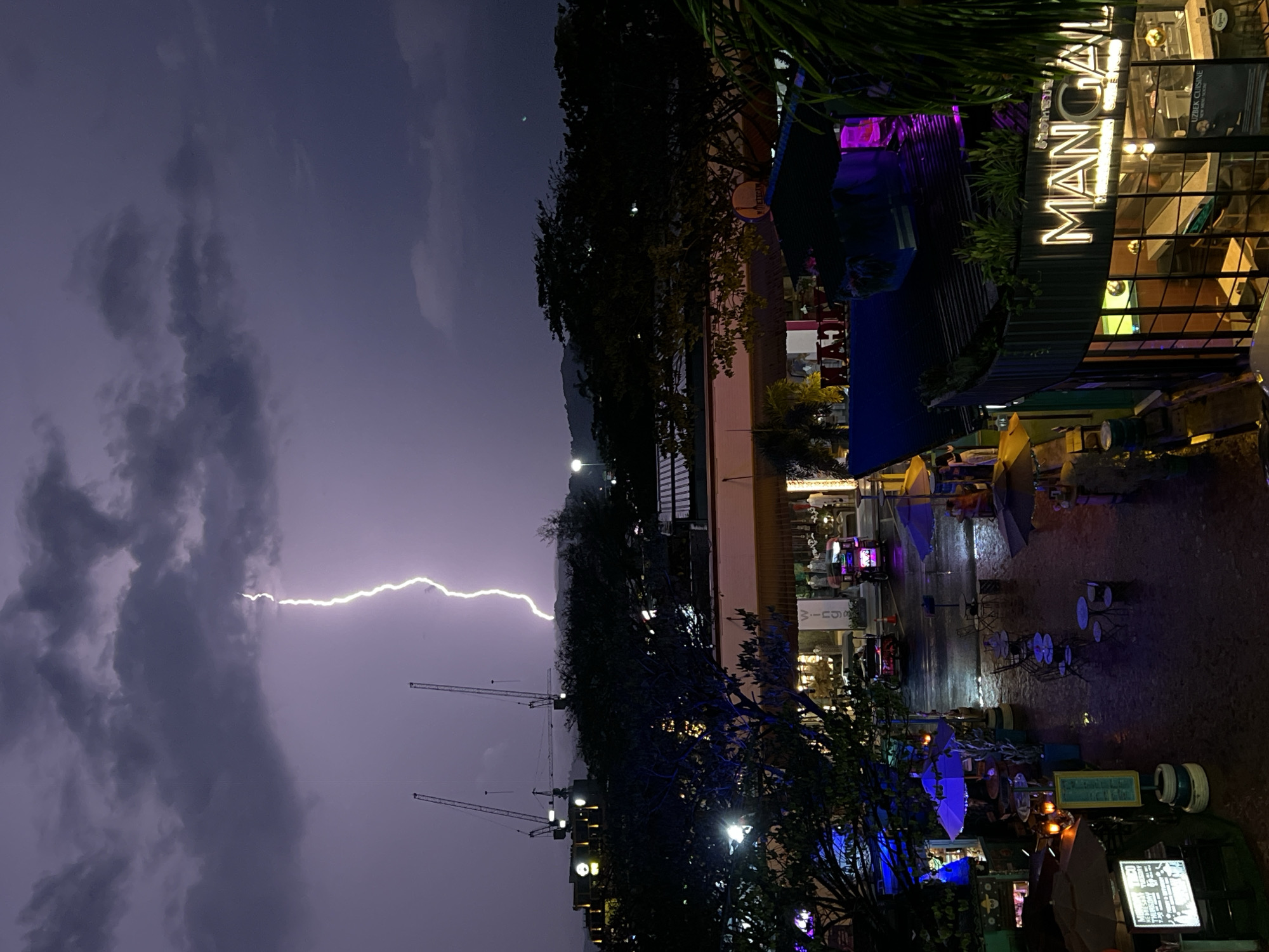 The image shows a cityscape during a thunderstorm at night. A prominent lightning strike illuminates the sky and the foreground buildings. The word "MANGAL" is visible in large, illuminated letters on a building. In the background are a few construction cranes. Streetlights and building lights reflect off wet surfaces below, suggesting rain is falling. Umbrellas are scattered around, indicating people have sought shelter from the downpour. The location is Unknown, Unknown.