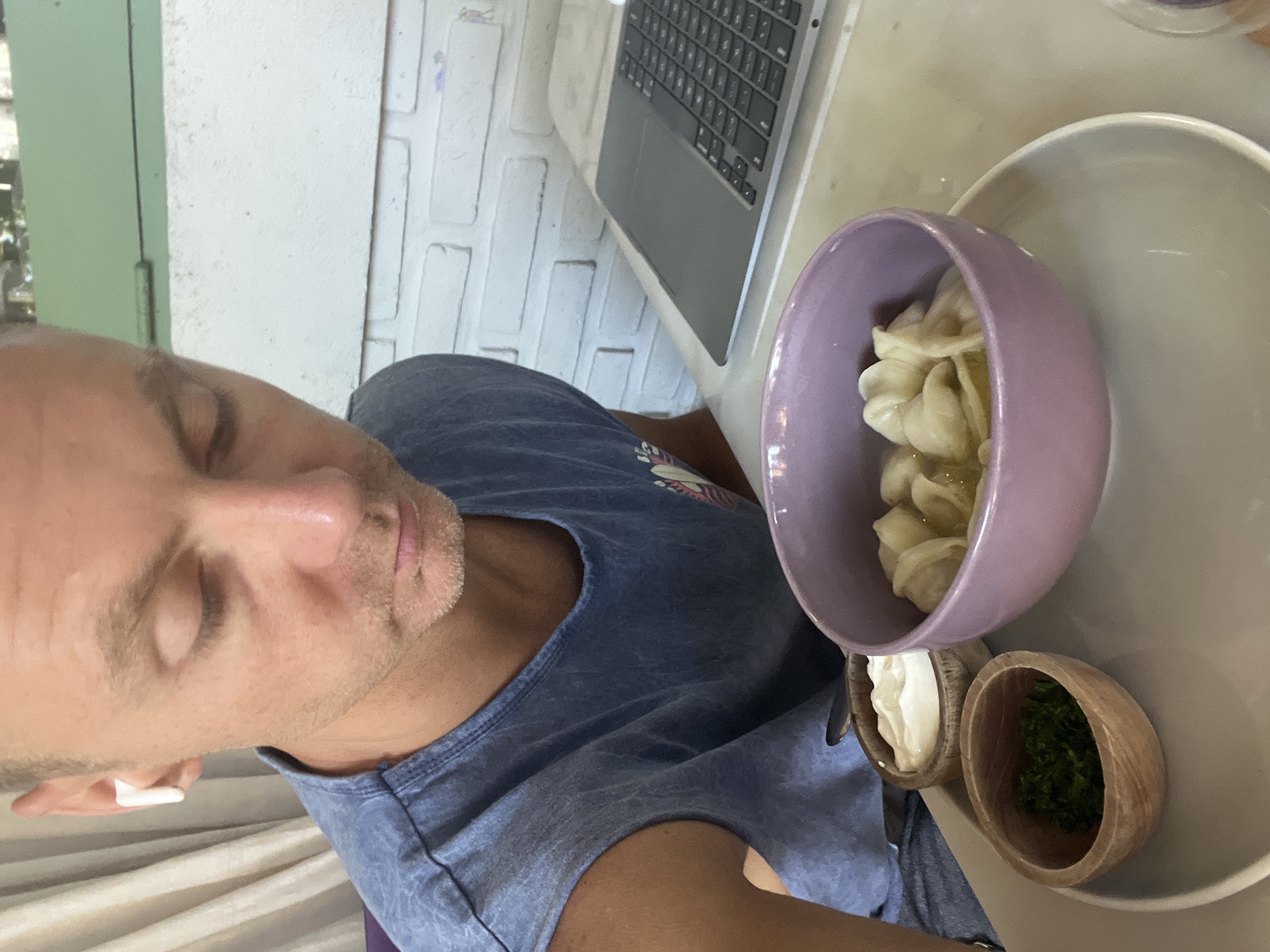 A close-up shot depicts a man sitting at a table with a laptop and a meal consisting of dumplings in a purple bowl, accompanied by small wooden bowls containing a white sauce and chopped greens, placed on a grey plate. The setting is likely indoors, suggested by the presence of a white brick wall. The man is wearing a blue t-shirt and has a white earbud in his ear. There is no definitive visual information to confirm that the location is Sukawati, Indonesia.