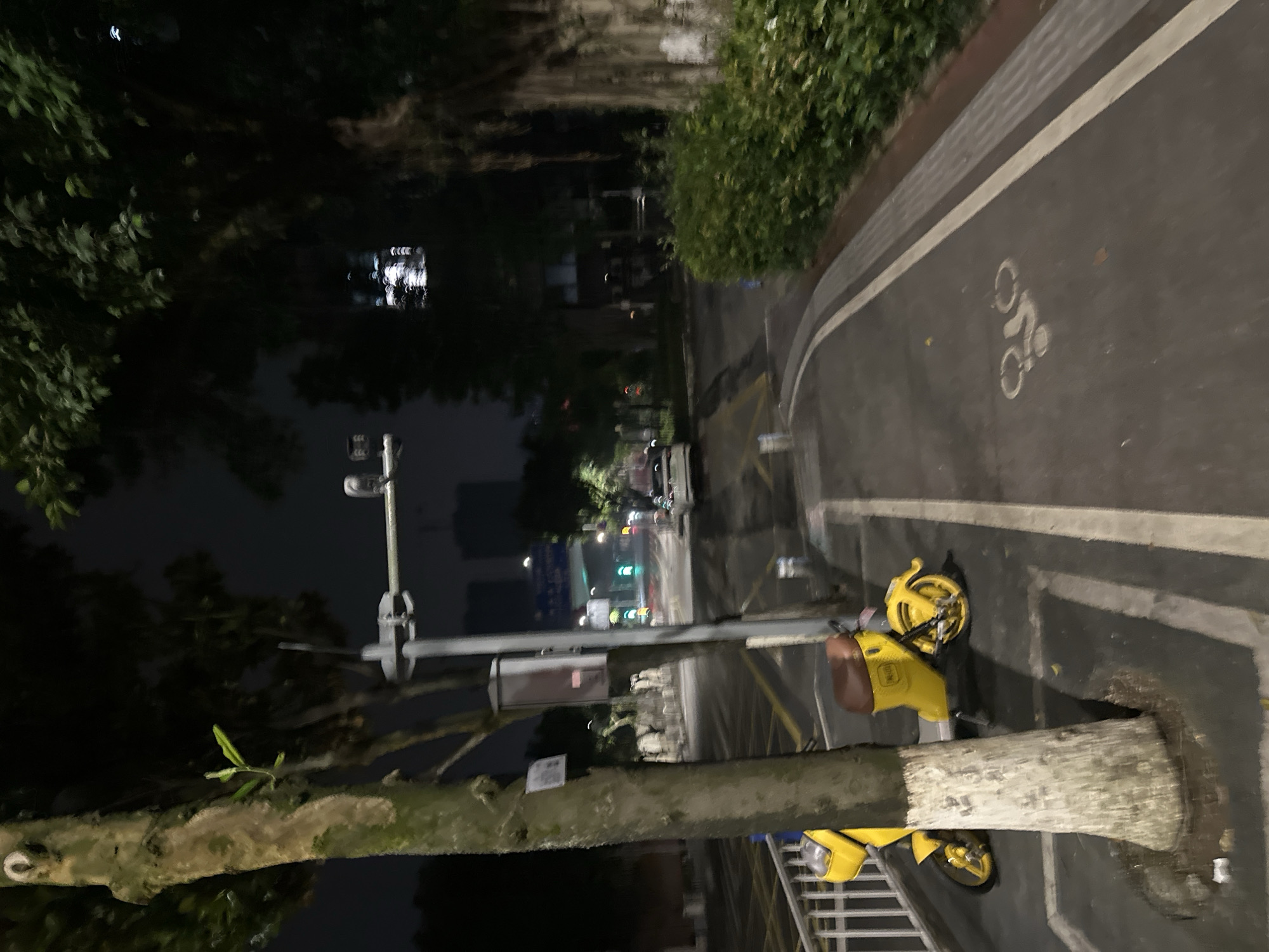 The image captures a nighttime scene on a city street in Chongqing High-tech Zone, China. A yellow electric scooter is parked next to a tree on the left side of the frame. The scooter has a brown seat and appears to be a shared mobility device. In the background, a road with traffic lights and vehicles is visible, with buildings and trees lining the street. A designated bicycle lane is marked on the asphalt with a white line and a bicycle symbol. The overall atmosphere is urban and the lighting suggests it is late at night.