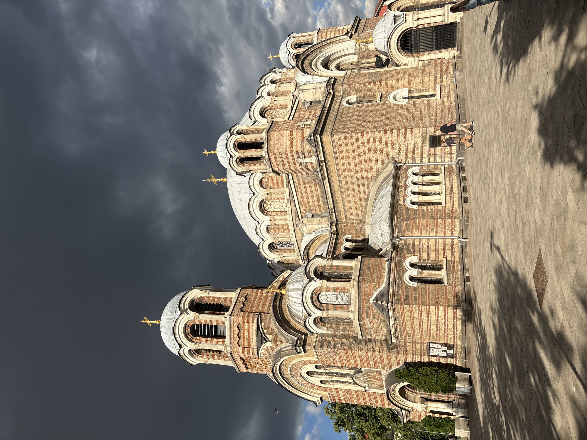 An exterior low-angle, full shot shows a large, ornate brick church with multiple domes and crosses against a dramatic, dark, cloudy sky. The church features arched windows and red and white striped brickwork. In the foreground, a paved courtyard is sparsely populated by three individuals. Two people, a man and a woman, walk from right to left across the courtyard. A third person, mostly obscured by shadow and viewed from behind, stands to the far right. Trees and shadows from unseen trees are visible in the courtyard.
